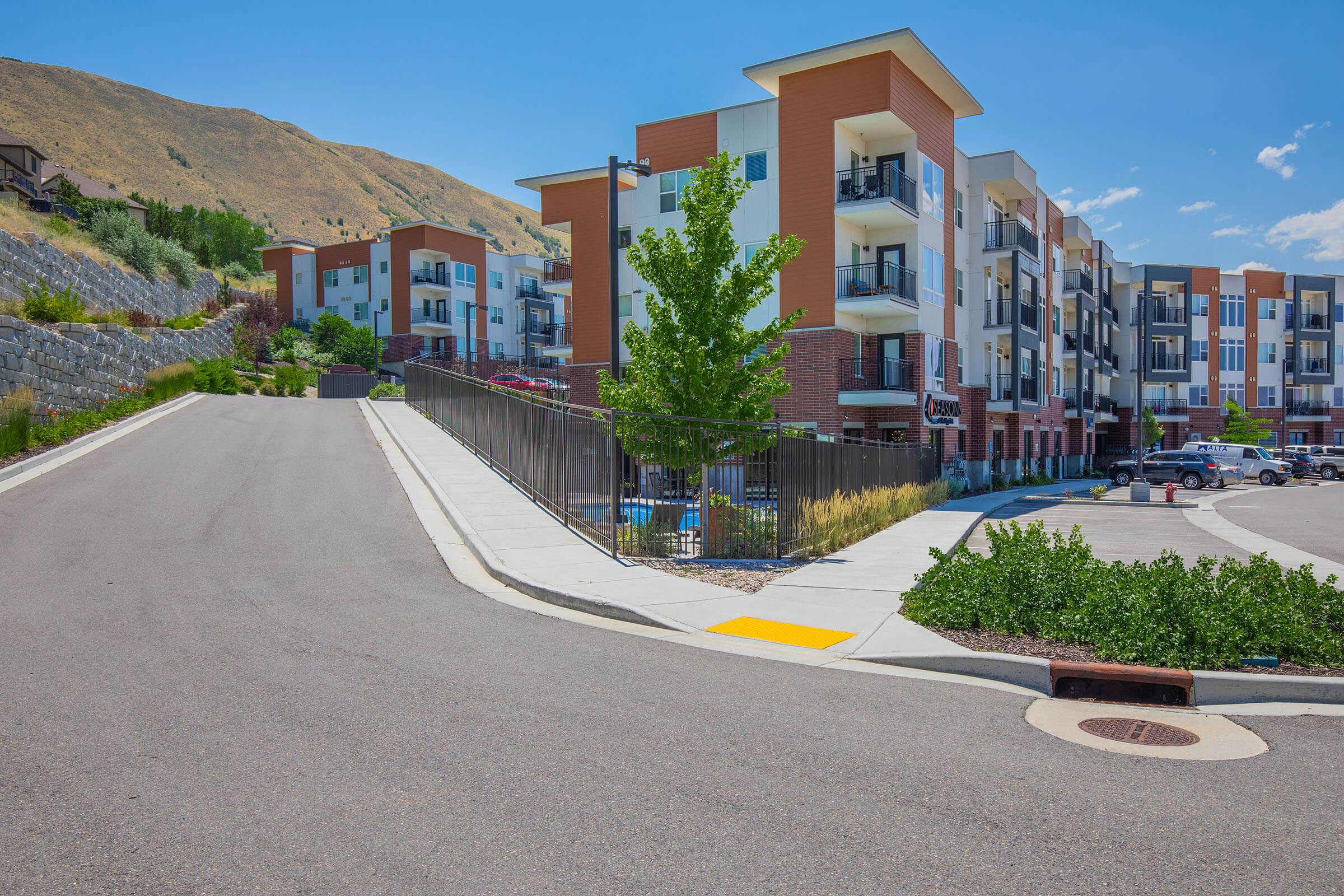 A modern apartment community is situated on a hillside, featuring multiple buildings with balconies. The foreground shows a paved road leading to the parking area, with landscaped greenery along the sidewalk. The sky is clear and blue, with a few clouds visible, and mountains in the background.