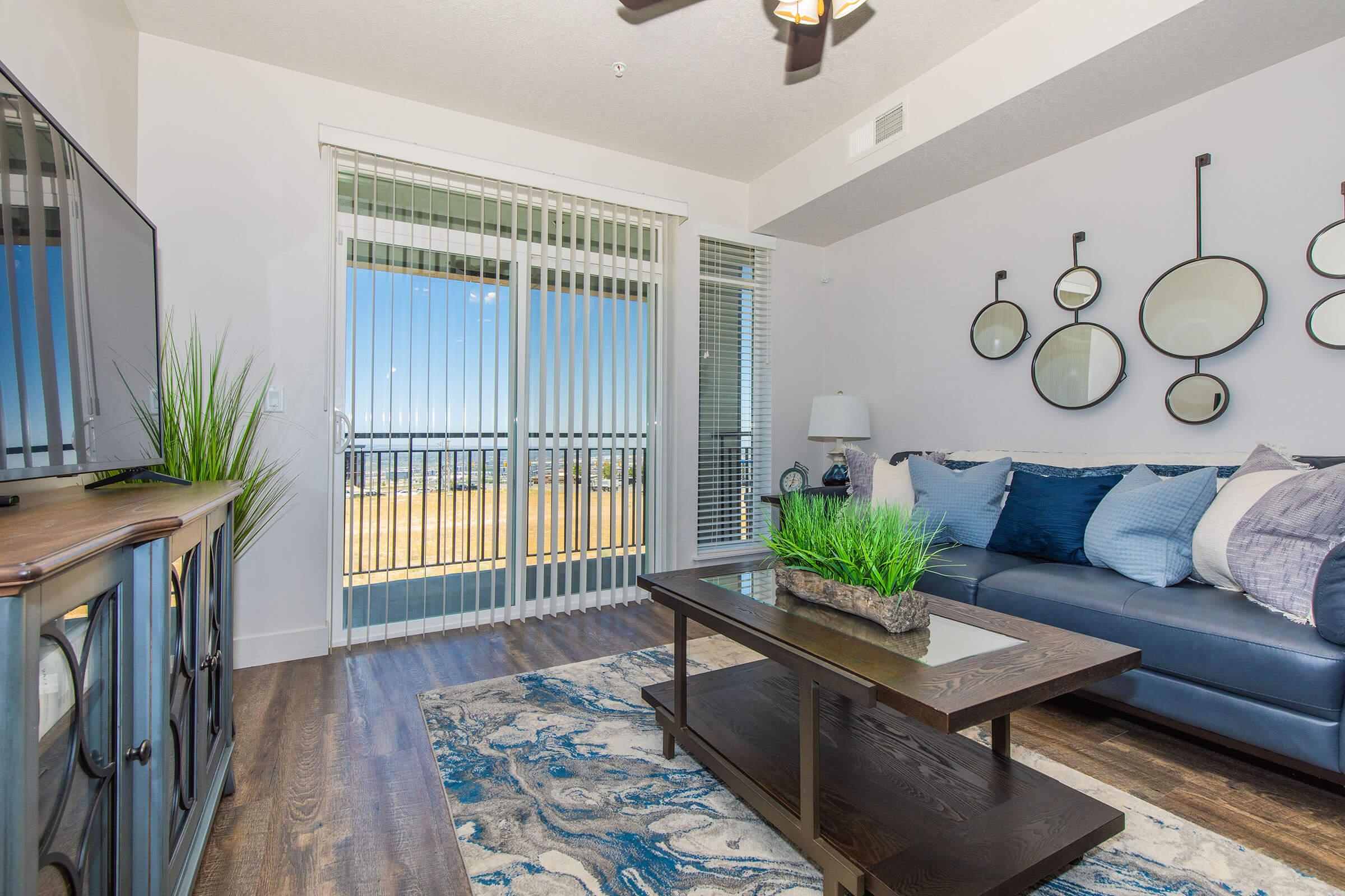 A modern living room featuring a navy blue sofa adorned with various decorative pillows. A stylish coffee table sits on a patterned area rug, surrounded by indoor plants. A TV on a wooden console faces sliding glass doors leading to a balcony, with natural light streaming in and circular mirrors on the wall.