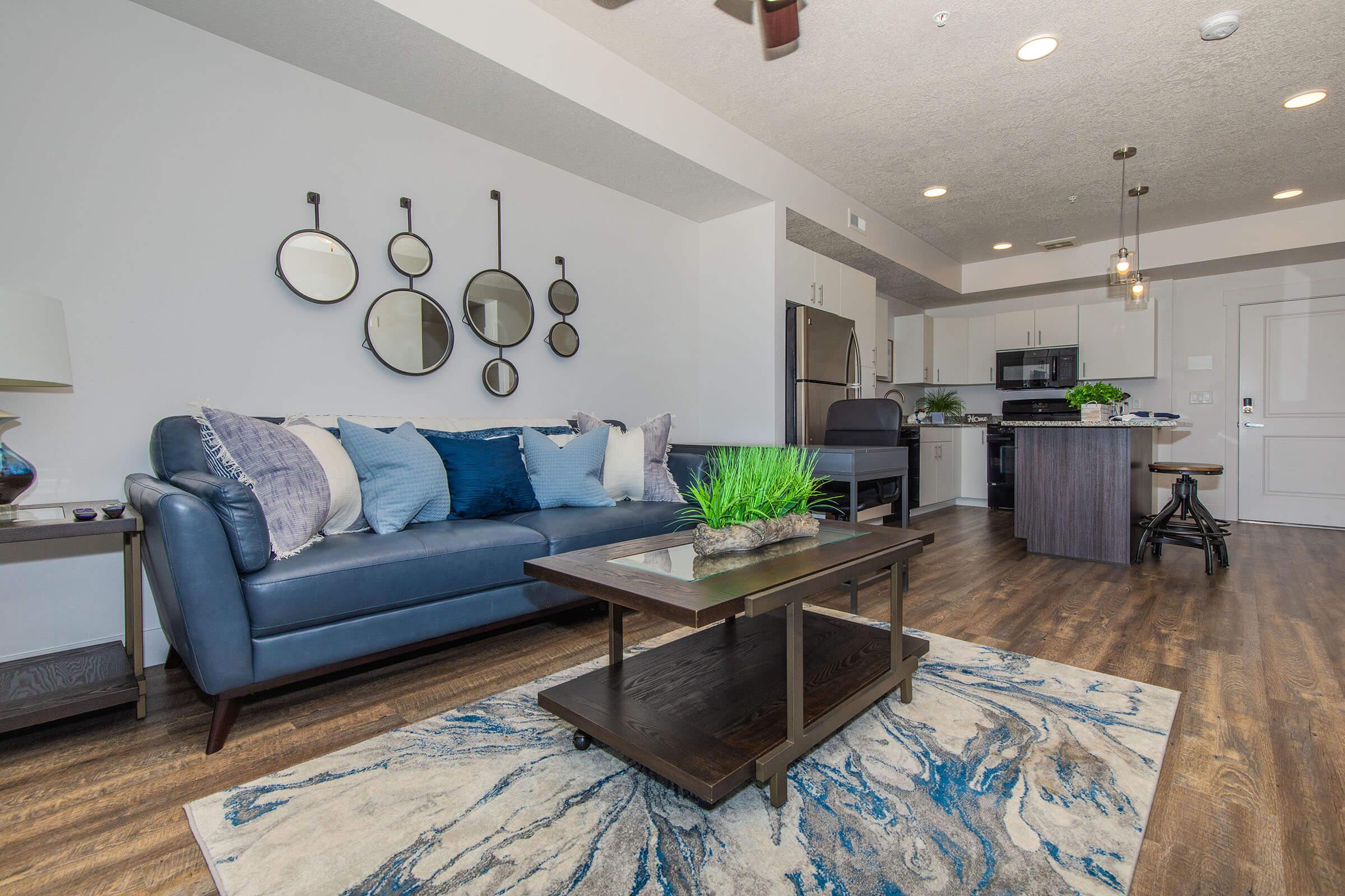 A modern living room featuring a blue couch adorned with various throw pillows, a stylish coffee table on a patterned area rug, and decorative mirrors on the wall. In the background, an open kitchen area with stainless steel appliances is visible, creating a cozy and inviting atmosphere.