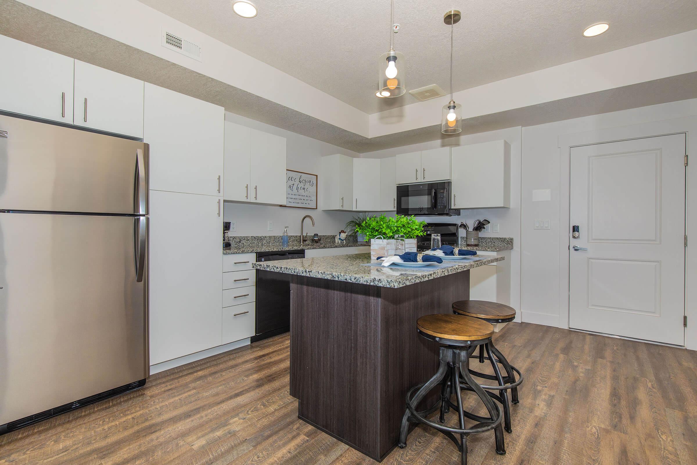 A modern kitchen with a central island featuring bar stools, stainless steel appliances, and sleek white cabinets. The countertop is made of granite, and a small plant adds a touch of greenery. The kitchen is well-lit with hanging light fixtures, creating a warm and inviting atmosphere.