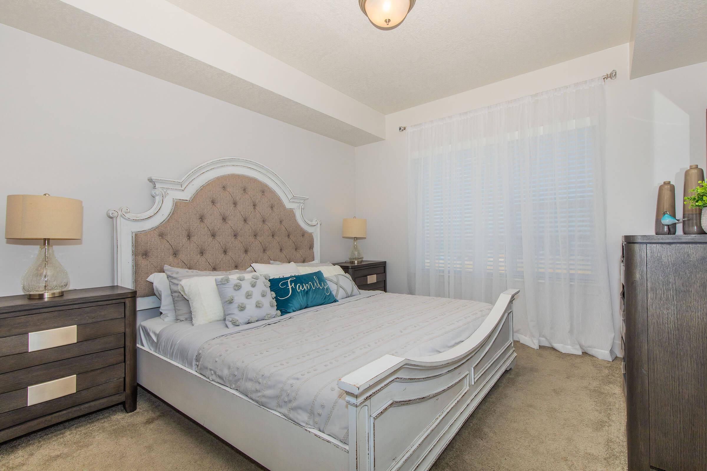 A well-decorated bedroom featuring a plush headboard bed with layered bedding and decorative pillows, including a blue "Family" pillow. There are two bedside lamps on dark wood nightstands, and large, sheer curtains letting in natural light. The room has neutral-colored walls and a cozy carpeted floor.