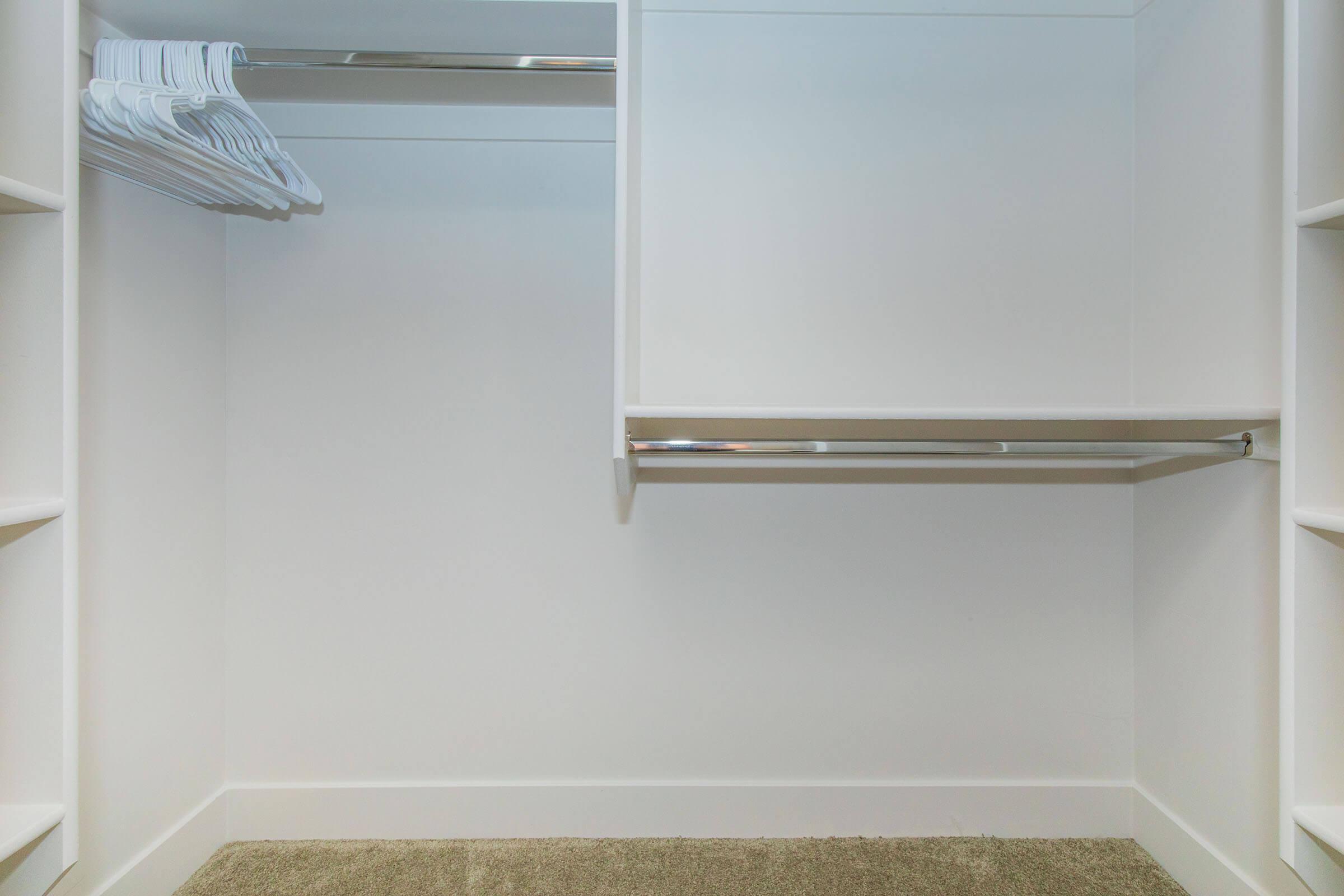 Empty closet interior with shelves on the left and a hanging rod on the right. There are a few white plastic hangers on the rod. The floor is covered with a light brown carpet, and the walls are painted white, creating a clean and minimalistic appearance.