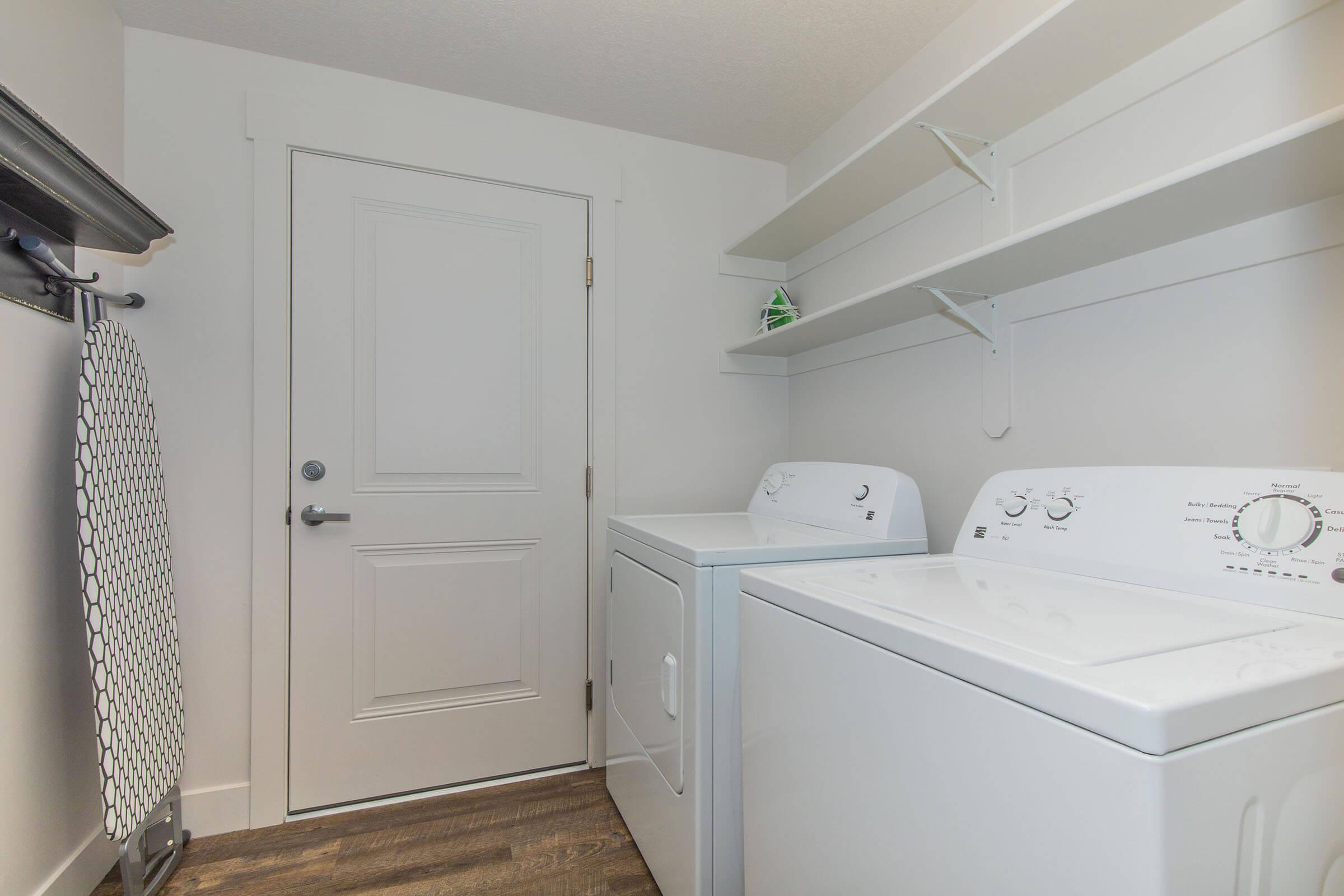 A clean laundry room featuring a white washing machine and dryer side by side, light-colored walls, and shelves above for storage. A door leads to another space, and there's a patterned towel hanging nearby. The flooring is wooden, adding warmth to the area.