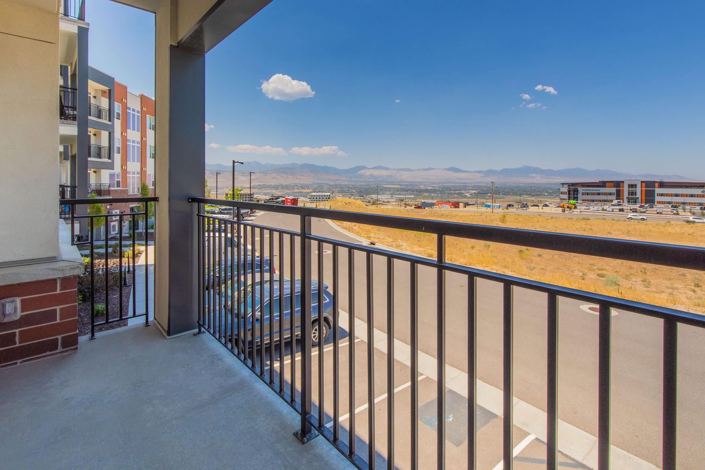 A balcony view overlooking a parking lot and a sprawling landscape with mountains in the distance. The sky is clear with a few clouds, and nearby buildings can be seen in the foreground. The scene captures a sunny day, highlighting the natural beauty of the surroundings.