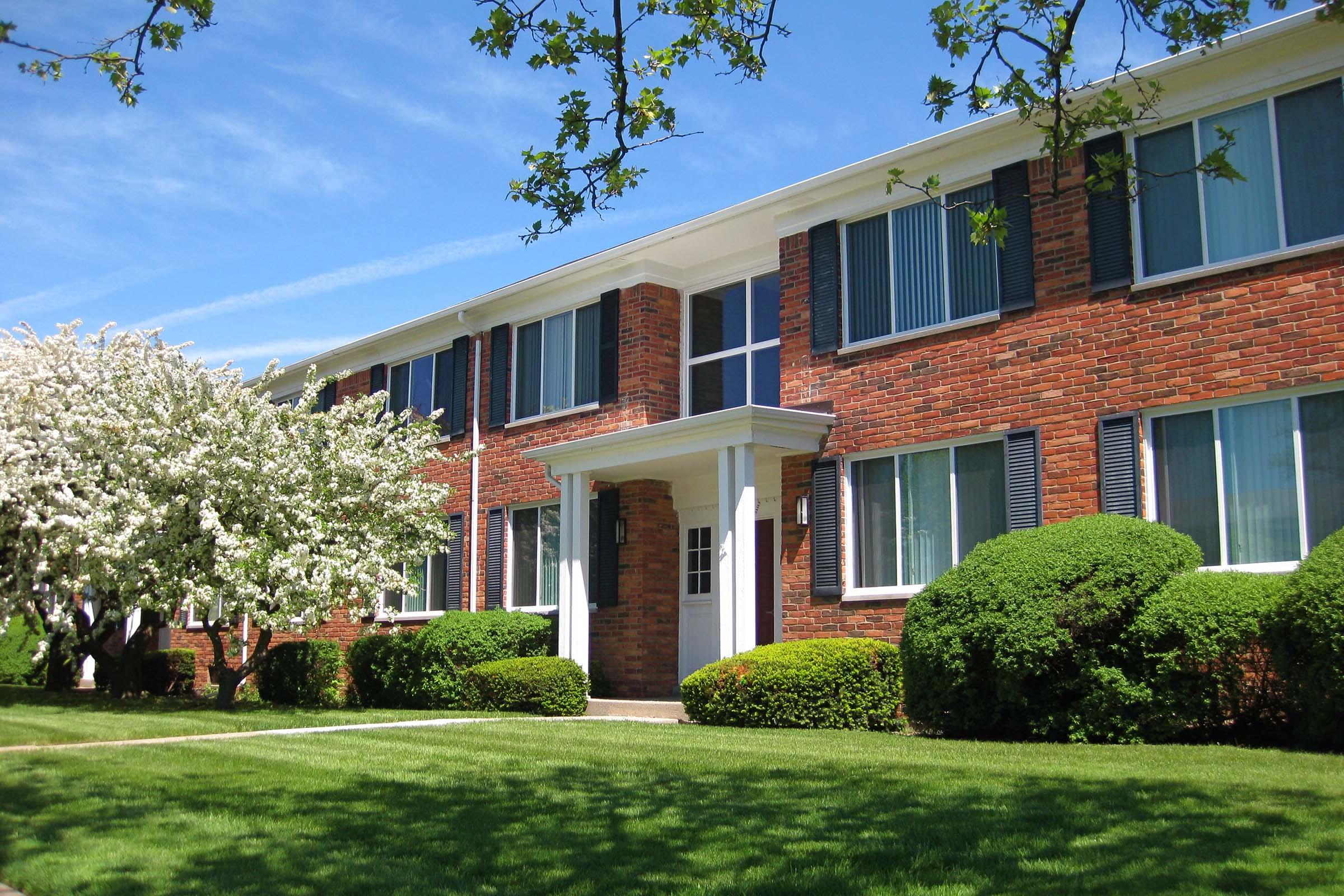 A brick apartment building with white trim, featuring large windows and a central entrance. In front, there is a well-maintained lawn with neatly trimmed bushes and a flowering tree, set against a clear blue sky.