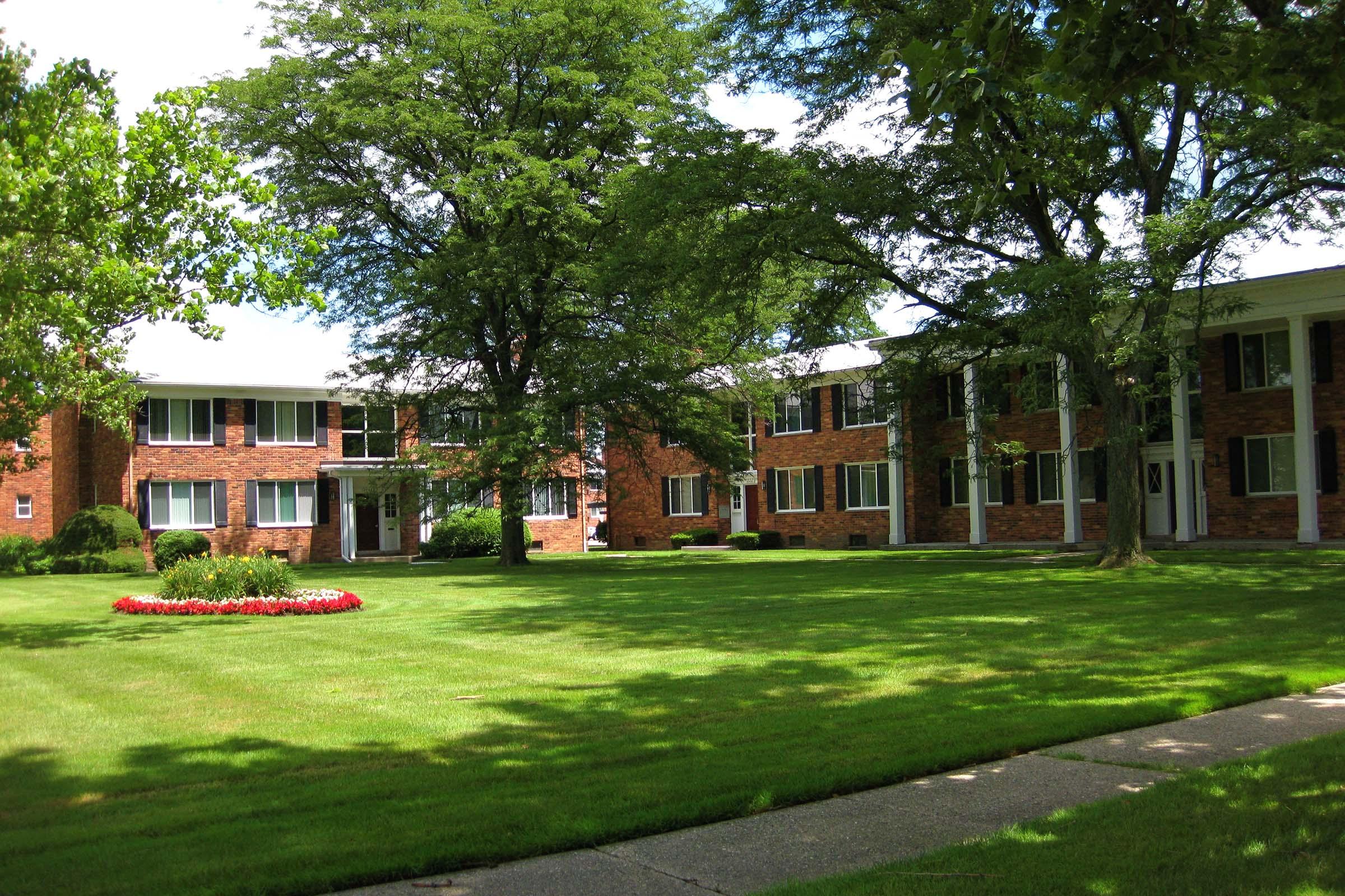 A well-maintained apartment complex featuring brick buildings surrounded by lush green grass and trees. The front lawn has a flower bed with red flowers, and a paved walkway leads up to the entrance of the buildings. Bright sunlight filters through the foliage, creating a welcoming atmosphere.