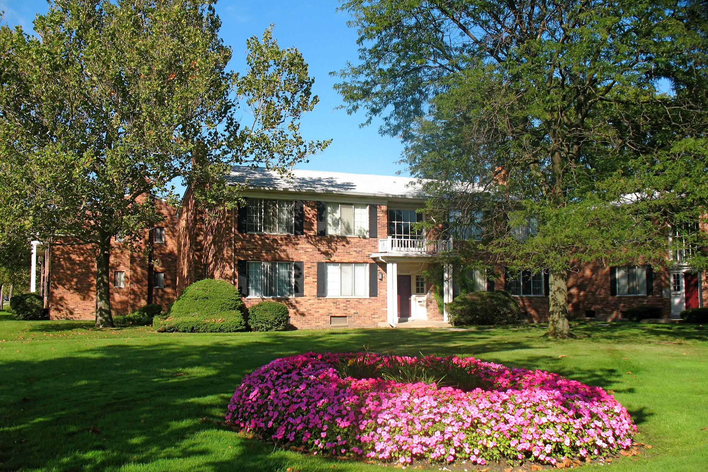 A two-story brick apartment building surrounded by greenery and large trees. In the foreground, a vibrant circular flower bed filled with colorful pink flowers contrasts with the well-manicured lawn. The sky is clear and blue, creating a bright, inviting atmosphere.