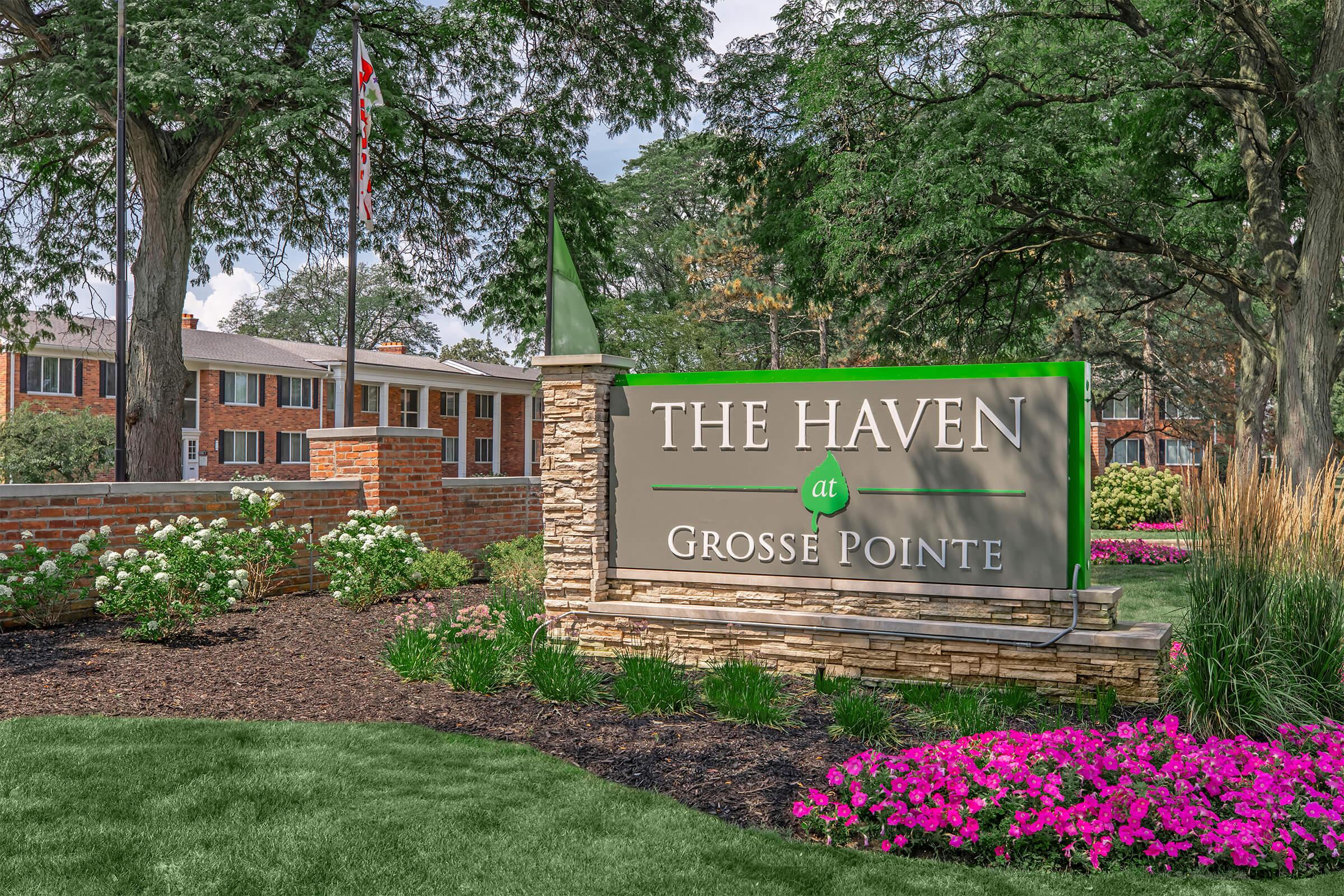 Sign for "The Haven at Grosse Pointe," featuring a green leaf design. Surrounding landscaping includes vibrant pink flowers and white blooms, with well-maintained grass and trees in the background. Buildings in the distance are modern and part of the complex.