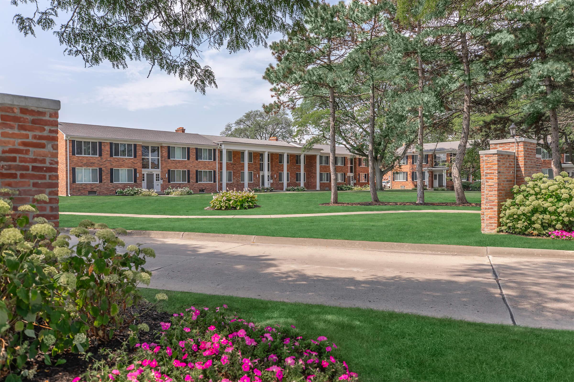 A well-maintained brick building with multiple windows, surrounded by green lawns and colorful flower beds featuring pink blooms. Tall trees provide shade in the background, and a paved path leads towards the entrance of the building. The scene conveys a peaceful and welcoming atmosphere.