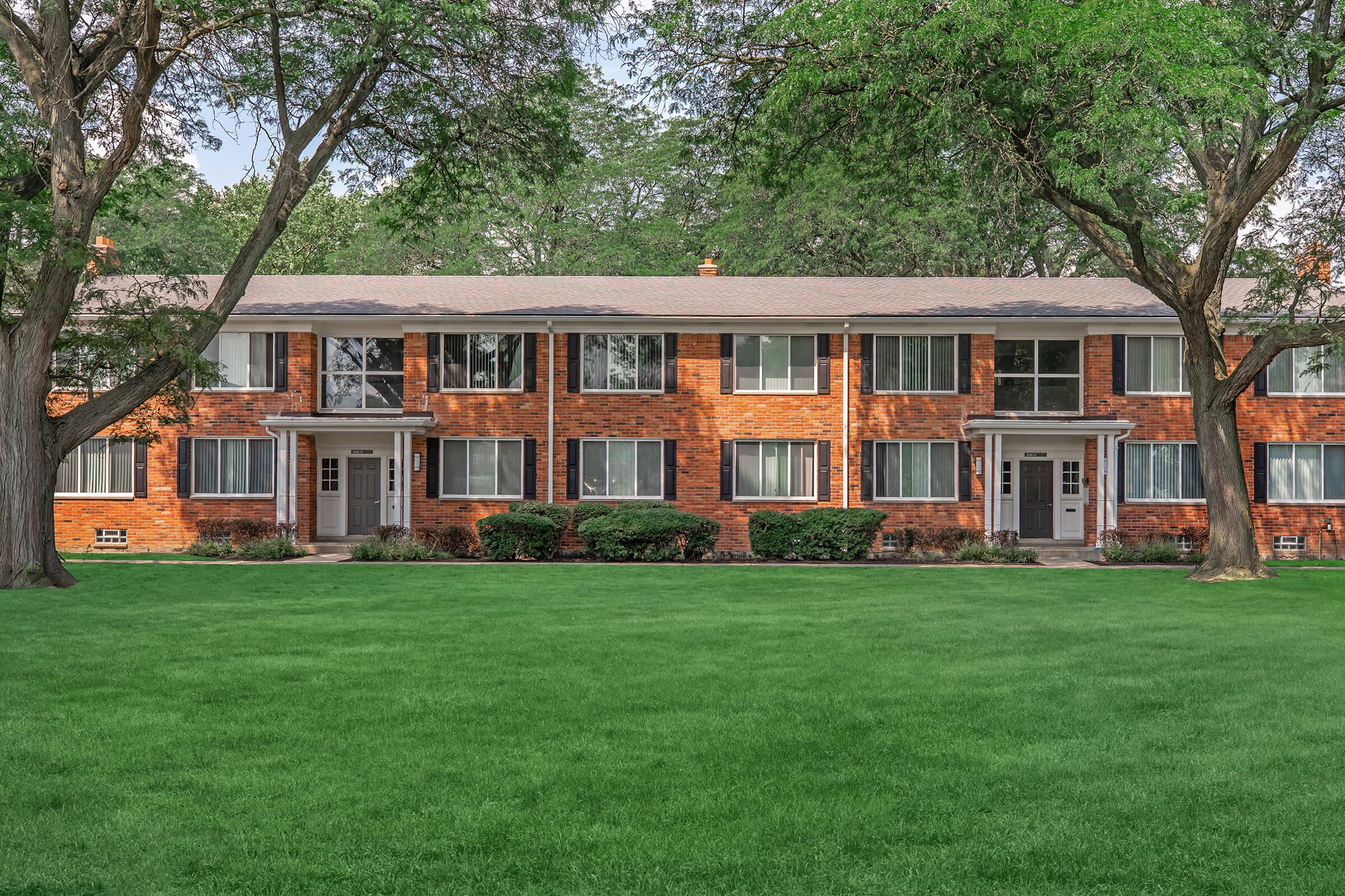 A two-story brick apartment building surrounded by lush green grass and trees. The building features multiple windows and front doors on each side, with a well-maintained lawn in front. Natural light highlights the building's warm tones.