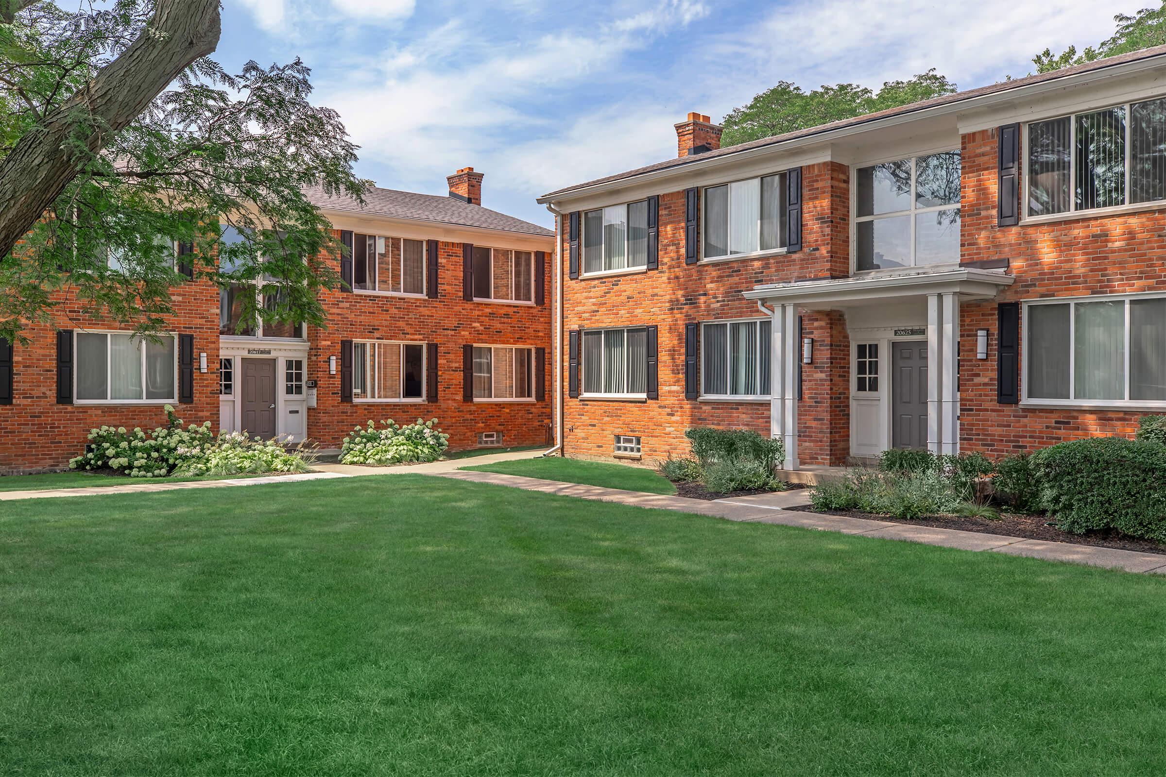 Two brick apartment buildings facing each other across a neatly maintained green lawn. Each building features large windows, white trim, and walkways leading to the entrances. Lush green shrubs and trees add to the pleasant, residential atmosphere on a sunny day.