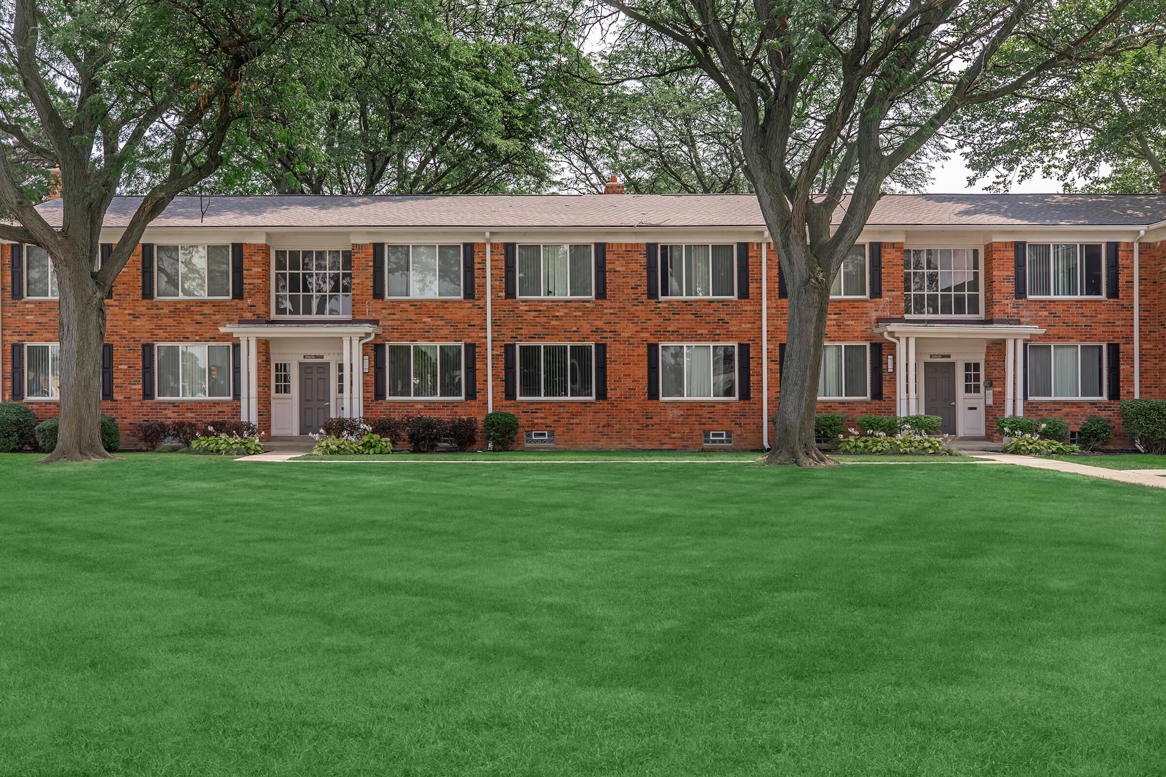 Two identical brick apartment buildings with white accents, surrounded by lush green grass and trees. The buildings feature multiple windows and entry doors, with well-maintained landscaping in front. The sky is bright and clear, enhancing the inviting atmosphere of the setting.