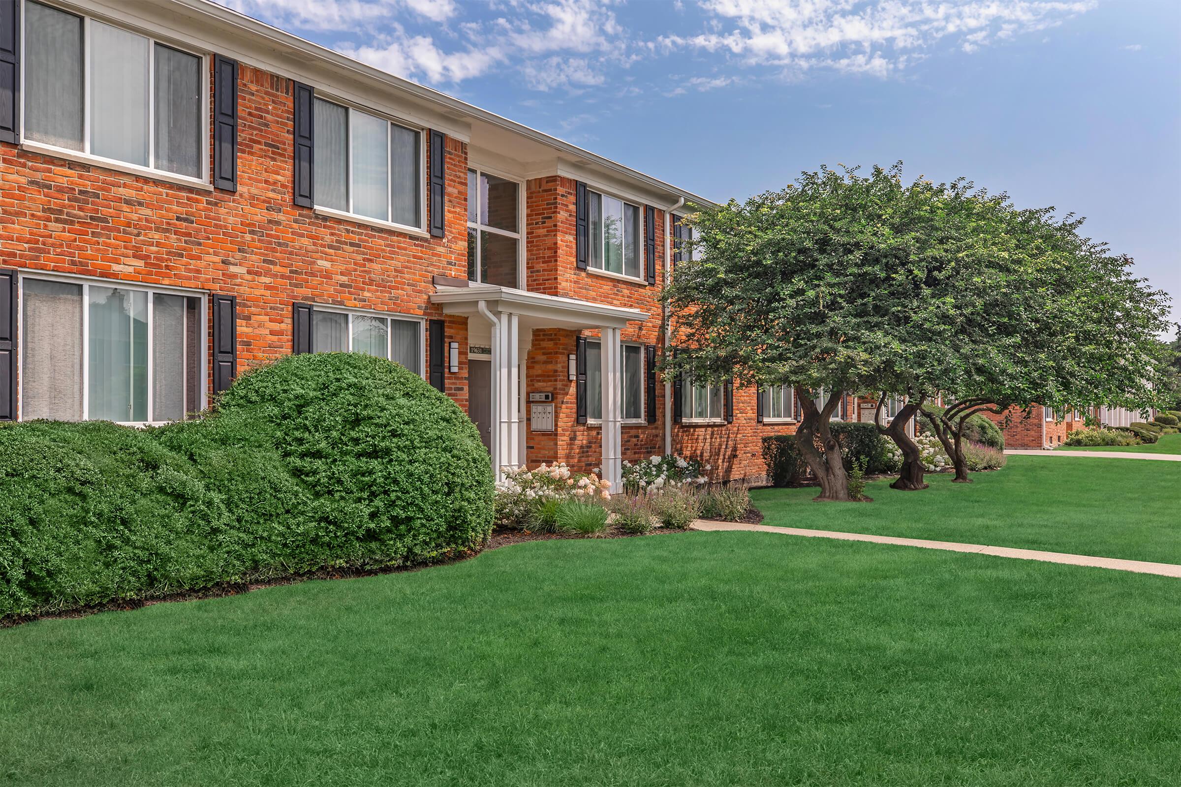 A brick apartment building with multiple windows is surrounded by neatly trimmed hedges and well-maintained green lawns. There are trees and flower beds adding to the landscaping, with a clear blue sky above. The setting gives a pleasant, welcoming feel to the residential area.