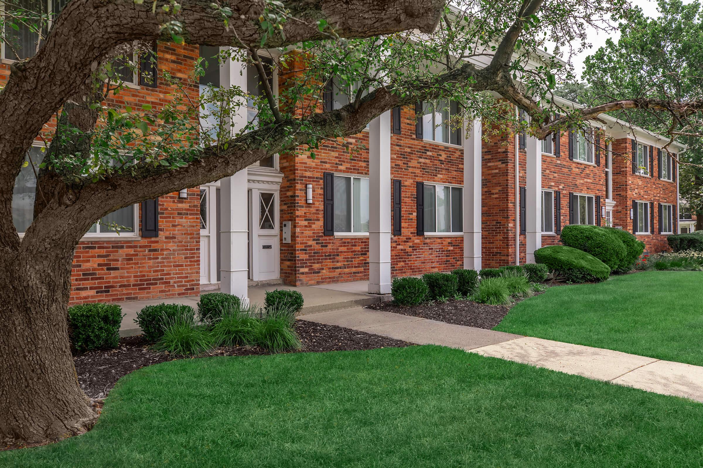 A well-maintained multi-unit brick apartment building with large windows and white columns. In the foreground, there is a manicured lawn with green grass and neatly trimmed bushes, while a mature tree with spreading branches stands nearby, providing shade to the entrance area.