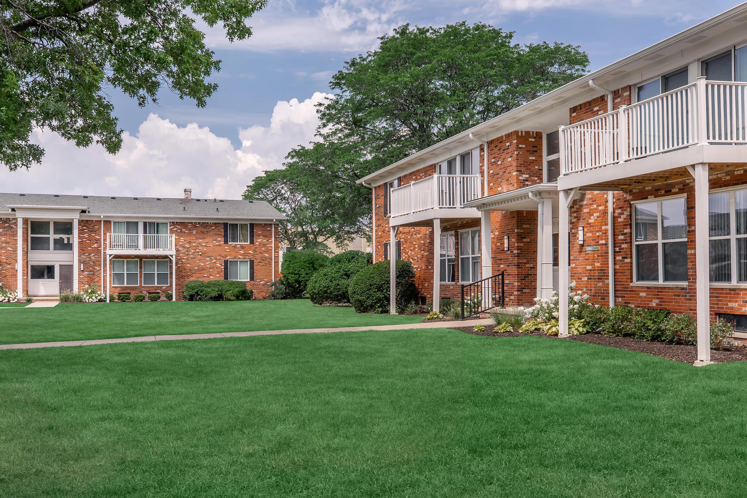 A well-maintained residential area featuring two brick apartment buildings, surrounded by lush green lawns and neatly trimmed bushes. The sky is partly cloudy, adding to the serene atmosphere. Balconies are visible on the building in the foreground, enhancing the inviting appearance of the community.