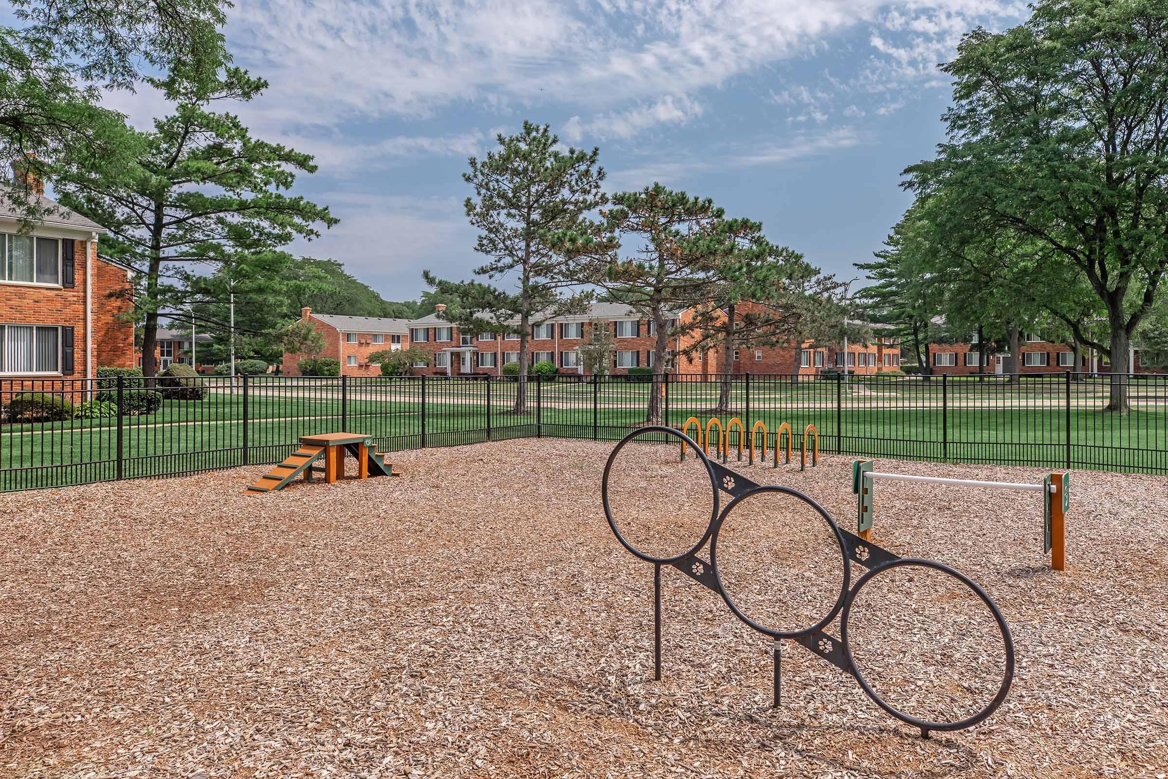 A playground area featuring various equipment, including climbing structures and a set of circular rings. The ground is covered with wood chips, and the playground is surrounded by a fence. In the background, several brick apartment buildings and trees are visible under a partly cloudy sky.