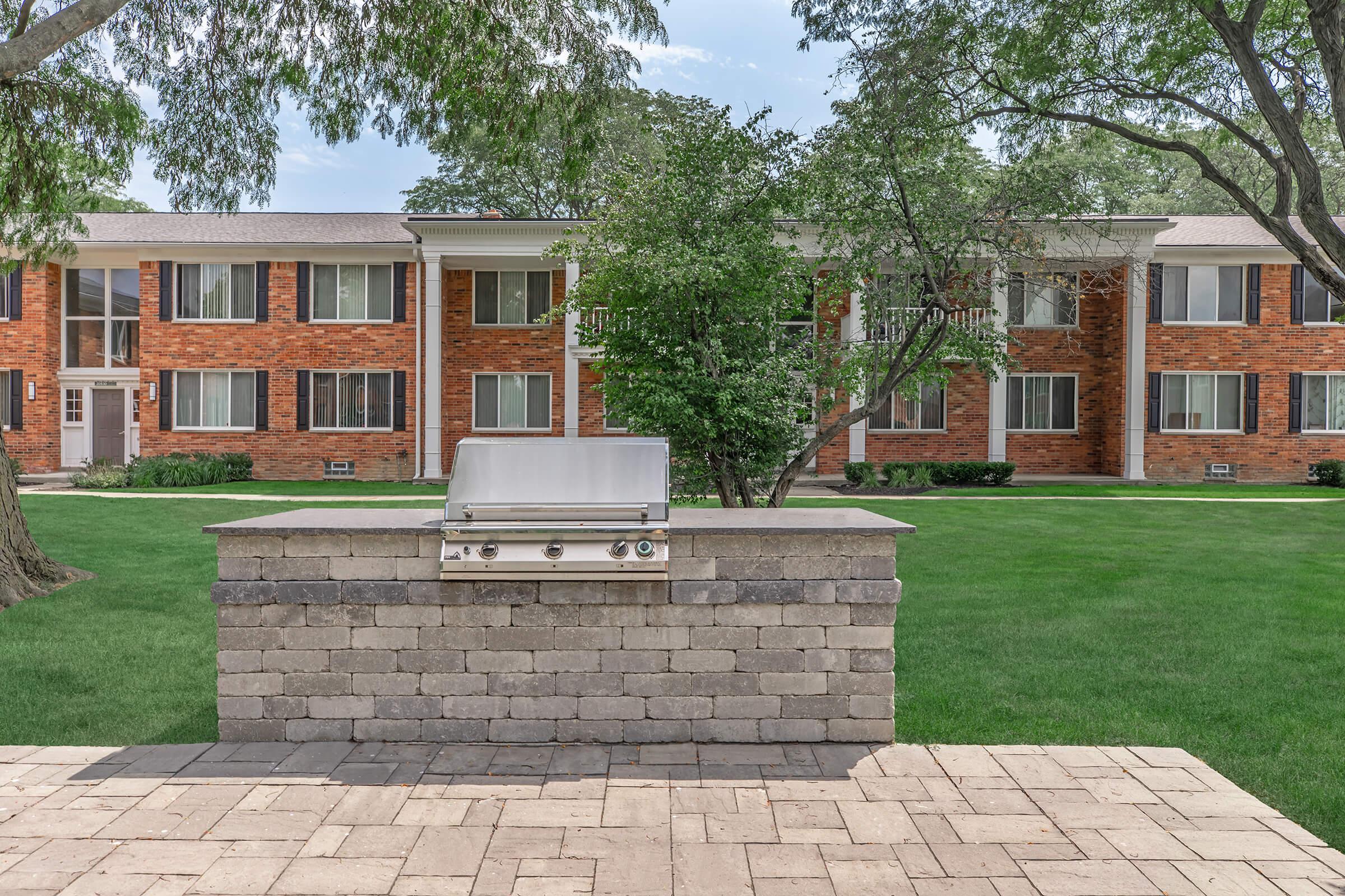 A modern outdoor grill set on a stone patio, surrounded by a lush green lawn and trees. In the background, there are several brick apartment buildings with multiple windows. The scene is bright and inviting, ideal for outdoor cooking and gatherings.