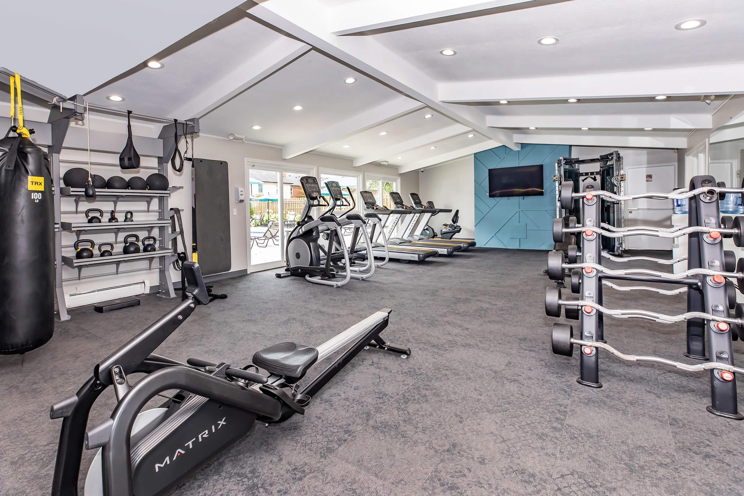 A modern gym interior featuring various exercise equipment, including cardio machines like treadmills and a rowing machine, weightlifting stations with free weights, a punching bag, and an interactive screen for workouts, all set in a well-lit, spacious environment.