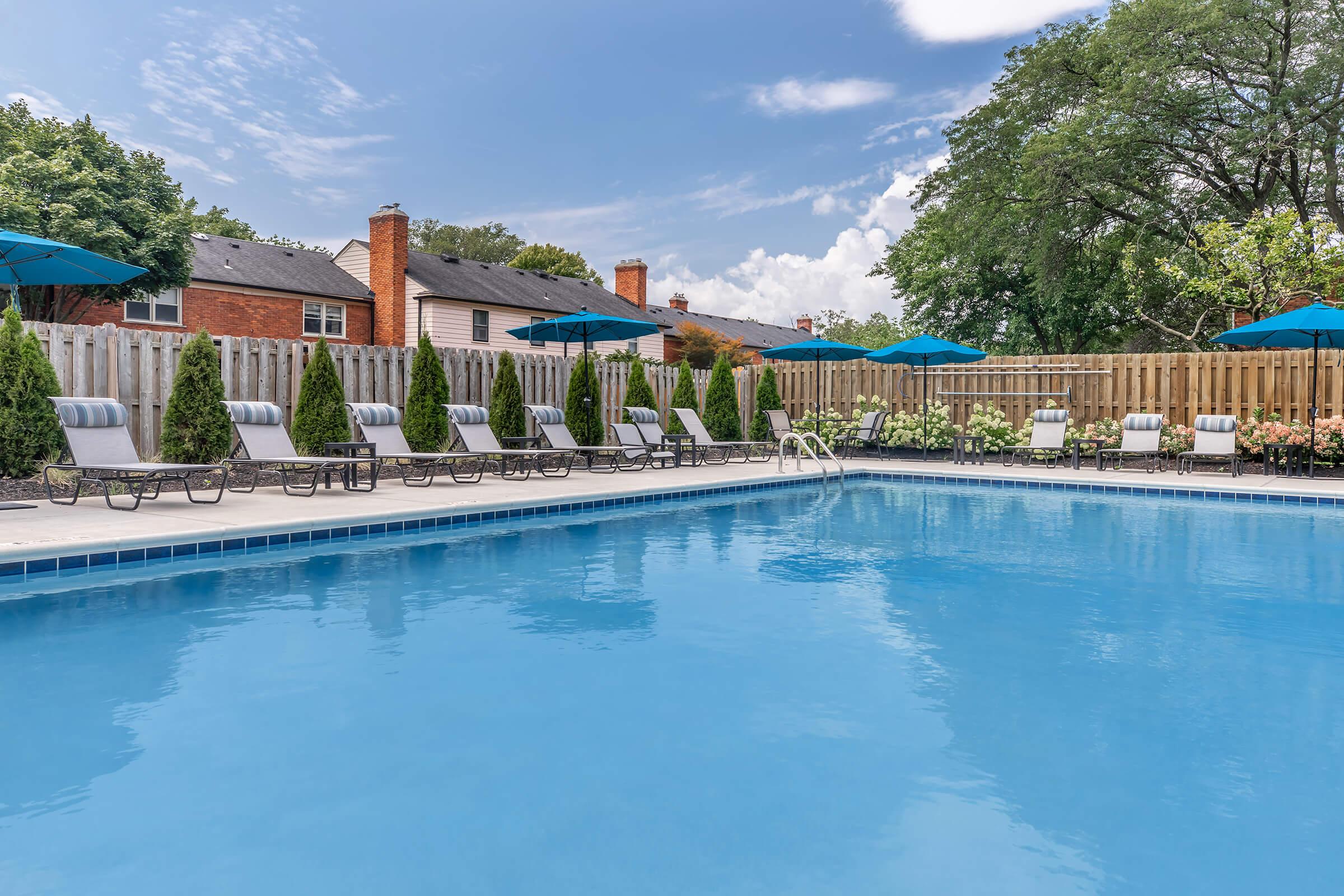 A clear blue swimming pool surrounded by lounge chairs and green umbrellas. Lush landscaping with neatly trimmed hedges and flower beds is visible along the pool's edge, set against a backdrop of residential buildings and a bright blue sky with scattered clouds.