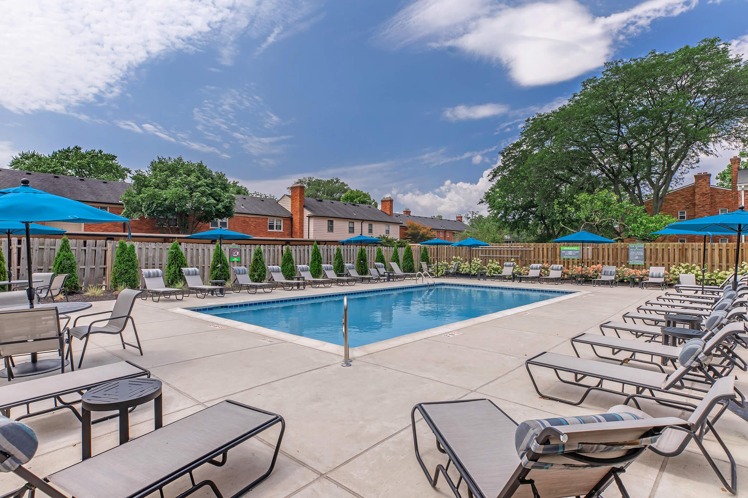 A swimming pool surrounded by lounge chairs and umbrellas on a bright day. The area features well-maintained landscaping with bushes and trees. Nearby buildings with red brick facades are visible in the background, and the sky is partly cloudy.