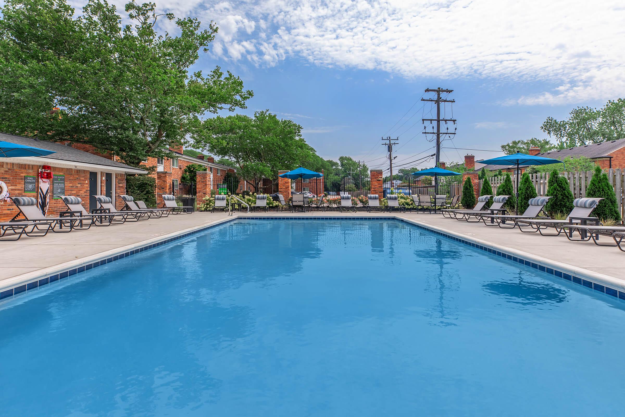 A clear blue swimming pool surrounded by lounge chairs and umbrellas. In the background, there are trees, a building with seating areas, and power lines. The sky is partly cloudy, creating a vibrant atmosphere for relaxation and leisure.
