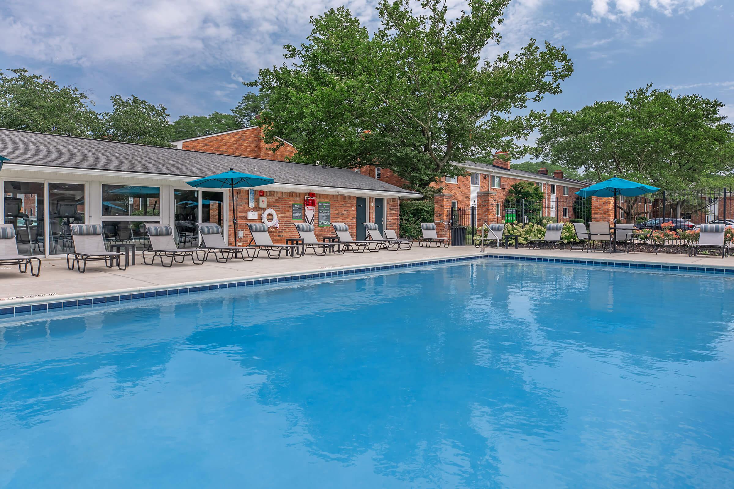 A clear blue swimming pool surrounded by lounge chairs and umbrellas. In the background, there are brick buildings and lush green trees, creating a serene atmosphere. The sky is partly cloudy, adding to the inviting scene.