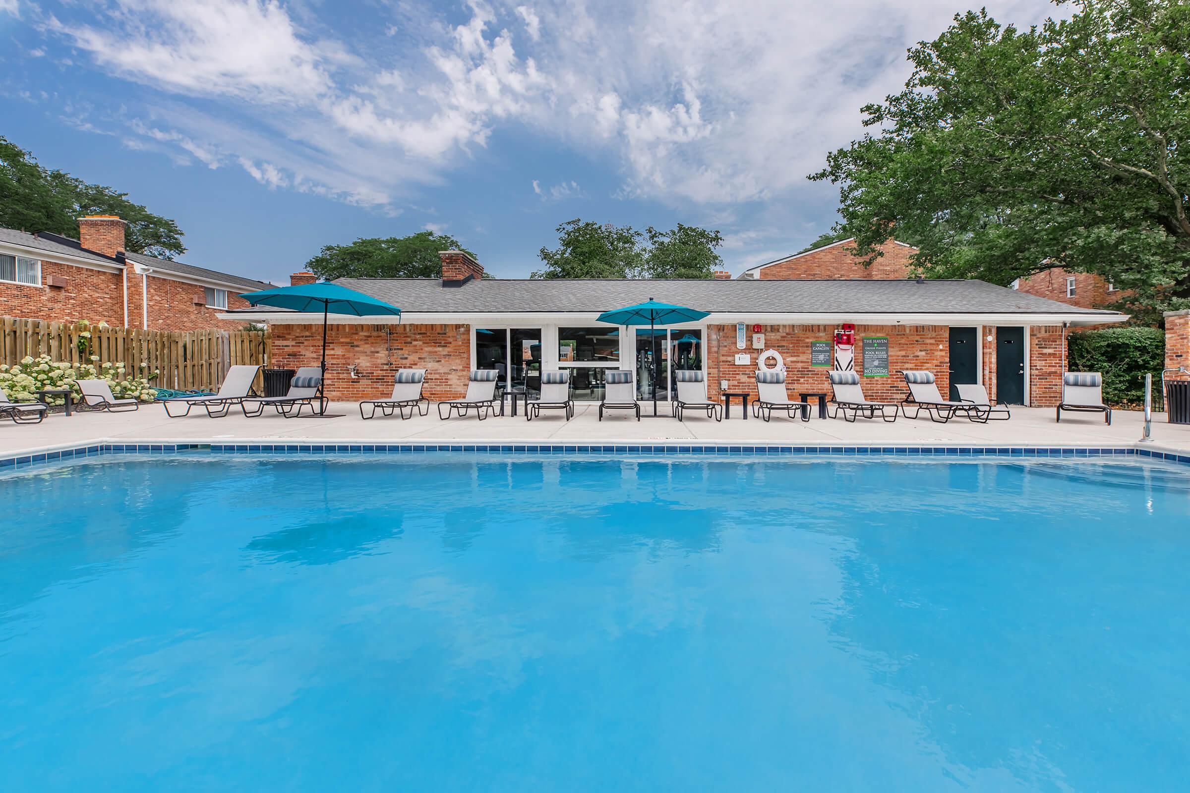 A sparkling blue swimming pool surrounded by lounge chairs and umbrellas, with a brick building in the background. The sky is bright with scattered clouds, and there is a wooden fence and greenery visible.