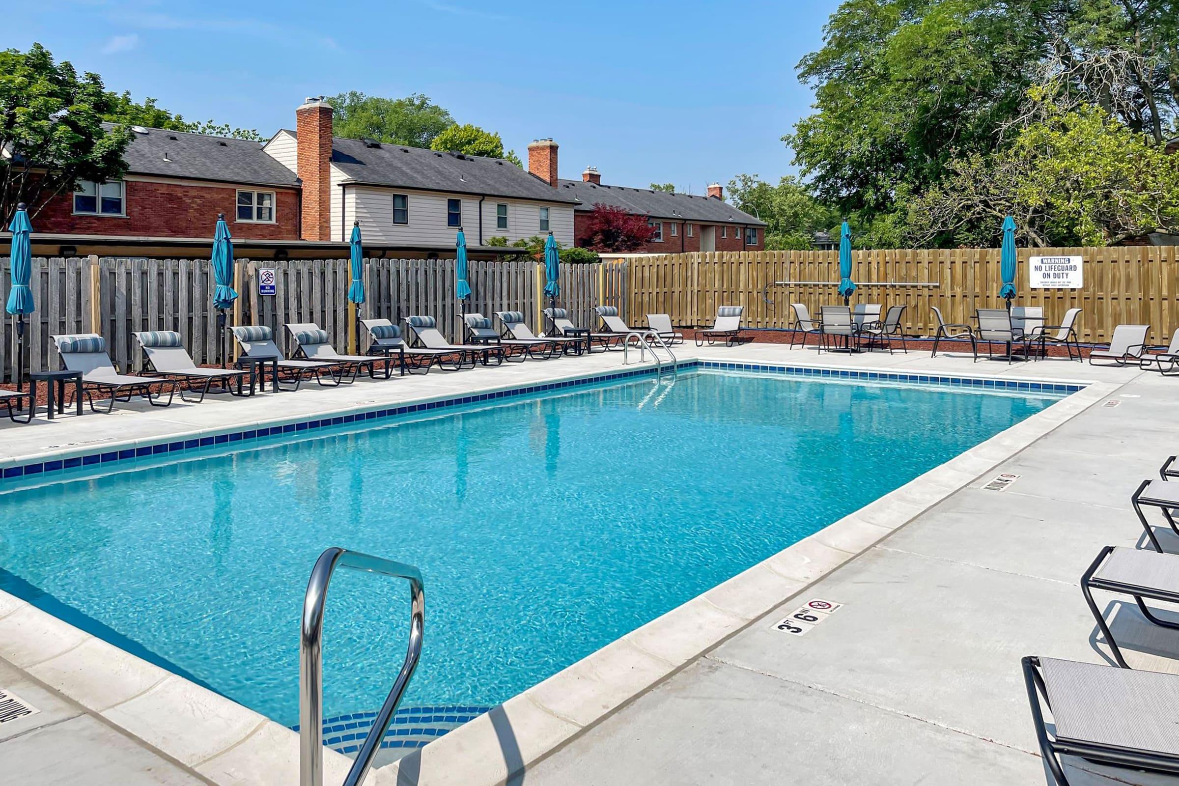 A clear swimming pool surrounded by lounge chairs and umbrellas. There are wooden fences on either side, with a few homes visible in the background. The sky is bright and clear, indicating a sunny day.