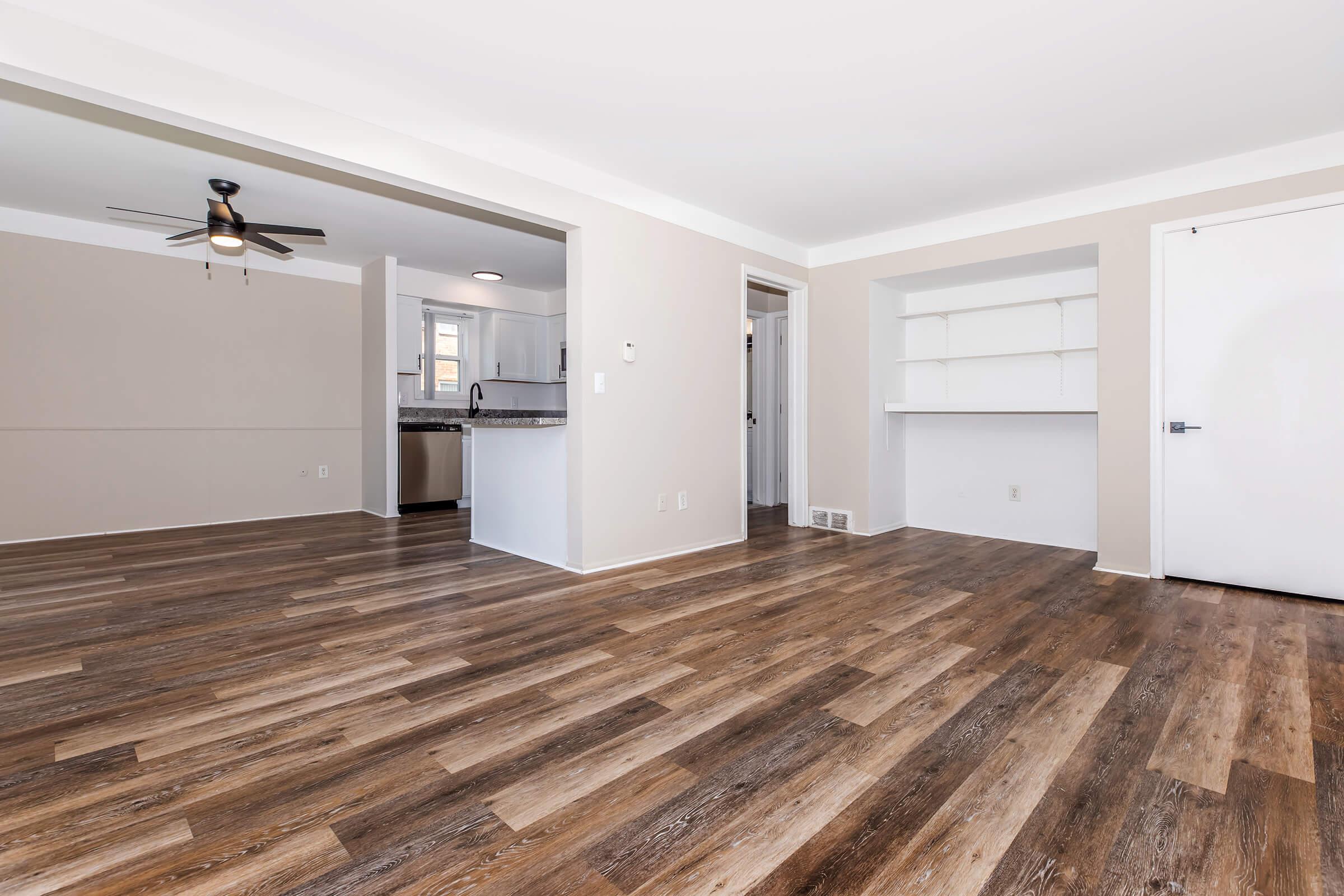 A spacious, well-lit living area featuring wood laminate flooring, a ceiling fan, and neutral-colored walls. To the left, an open passage leads to a modern kitchen with white cabinetry. On the right, a built-in shelf unit adds functionality. The room is empty, showcasing its potential for furniture placement.