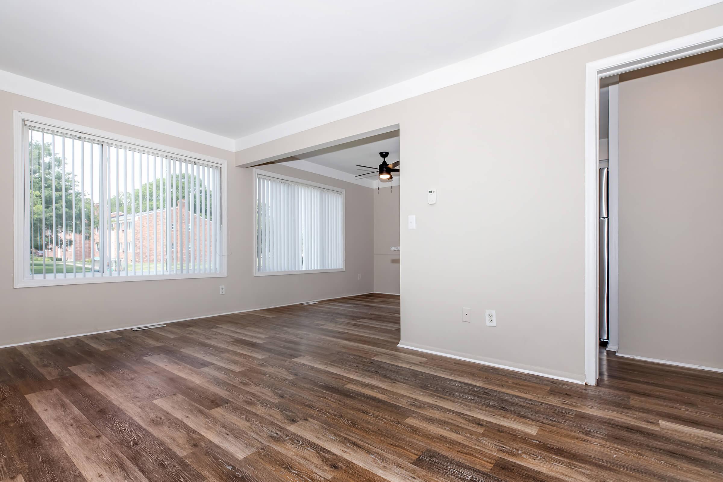 Bright and spacious living room with large windows featuring vertical blinds, wooden flooring, and a ceiling fan. The room has neutral wall colors and an open layout connecting to an adjacent area. Natural light fills the space, creating a warm and inviting atmosphere.