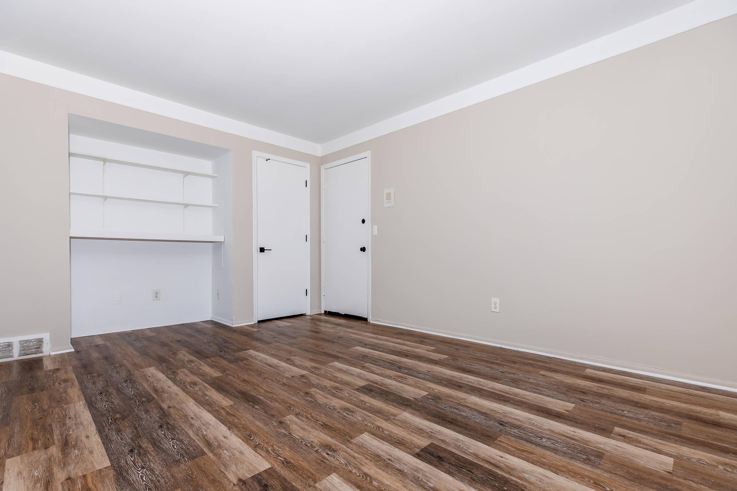 Empty room with light brown walls and wooden flooring. A small built-in shelf is visible on the left side, while two closed doors are on the right side. The room is well-lit and has a clean, minimalist appearance. No furniture or decorations are present.
