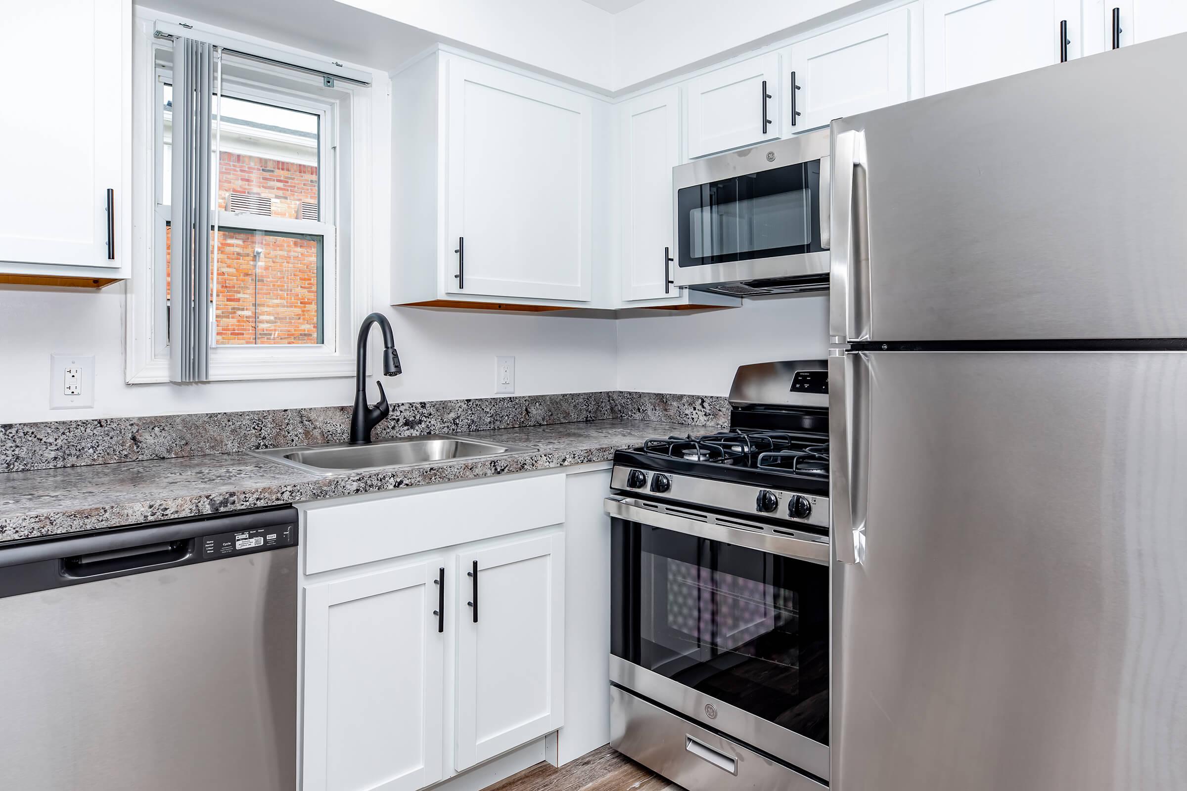 A modern kitchen featuring white cabinets, stainless steel appliances including a refrigerator, oven, and dishwasher, granite countertops, and a window that provides natural light. The design emphasizes a clean and contemporary aesthetic with a functional layout.