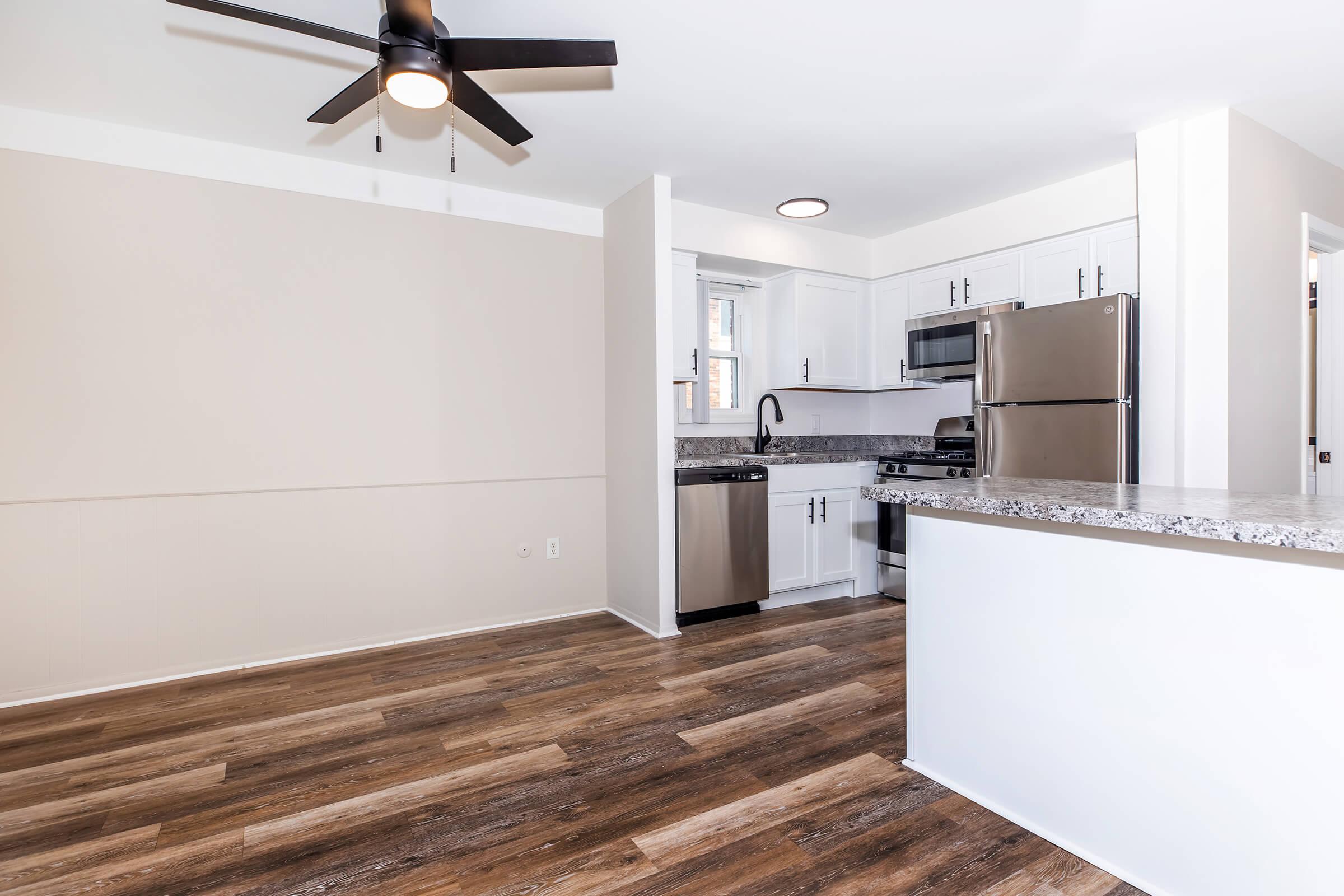A modern kitchen featuring white cabinets, stainless steel appliances, and a granite countertop. The space has a ceiling fan, wood-like flooring, and ample natural light coming through a window. The layout is open, connecting to a dining area with light-colored walls.