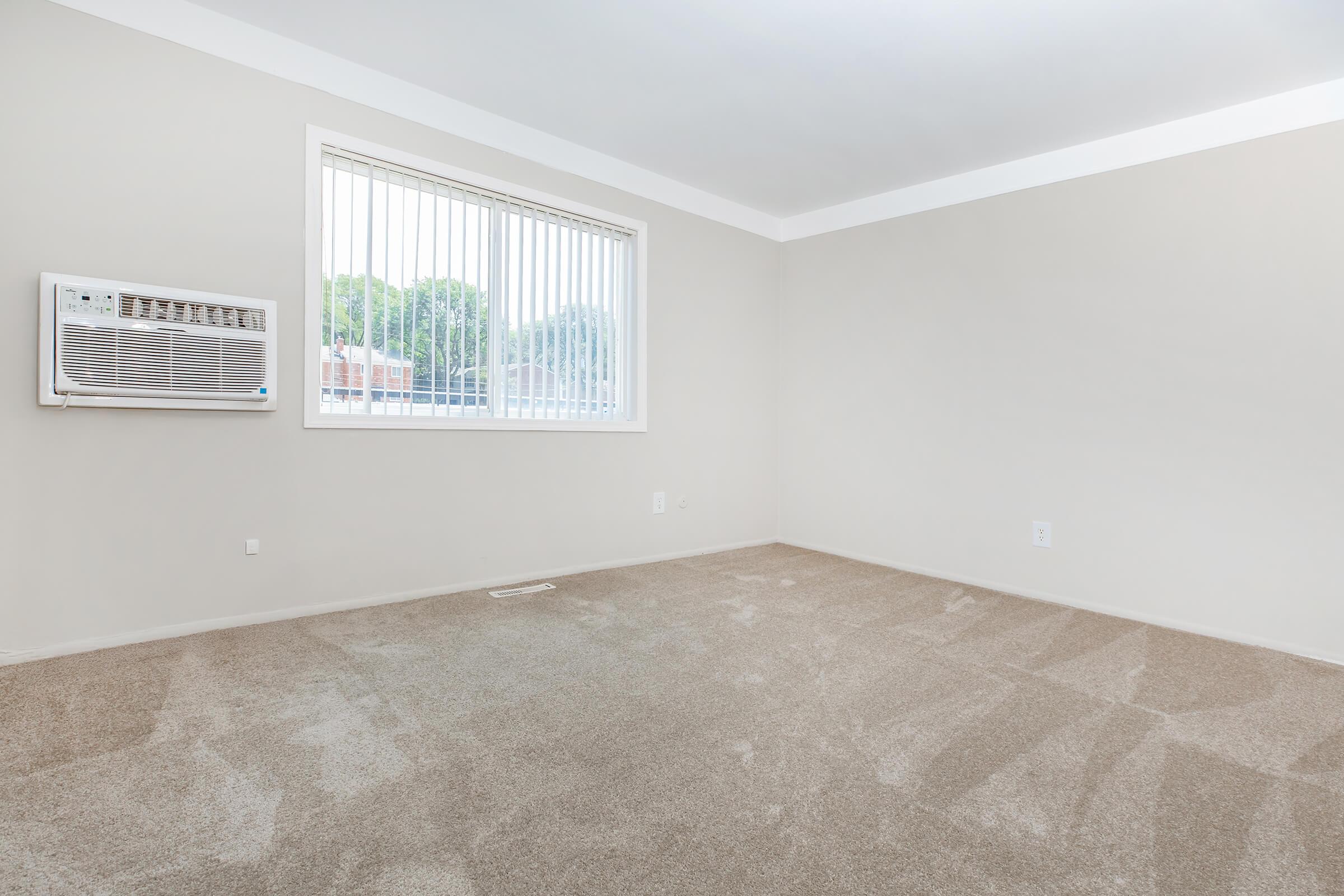 A bare, empty room with light beige walls and a large window covered by vertical blinds. An air conditioning unit is mounted on the wall next to the window, and the floor is carpeted in a light tan color. The room has no furniture or decorations, creating a spacious, minimalistic feel.