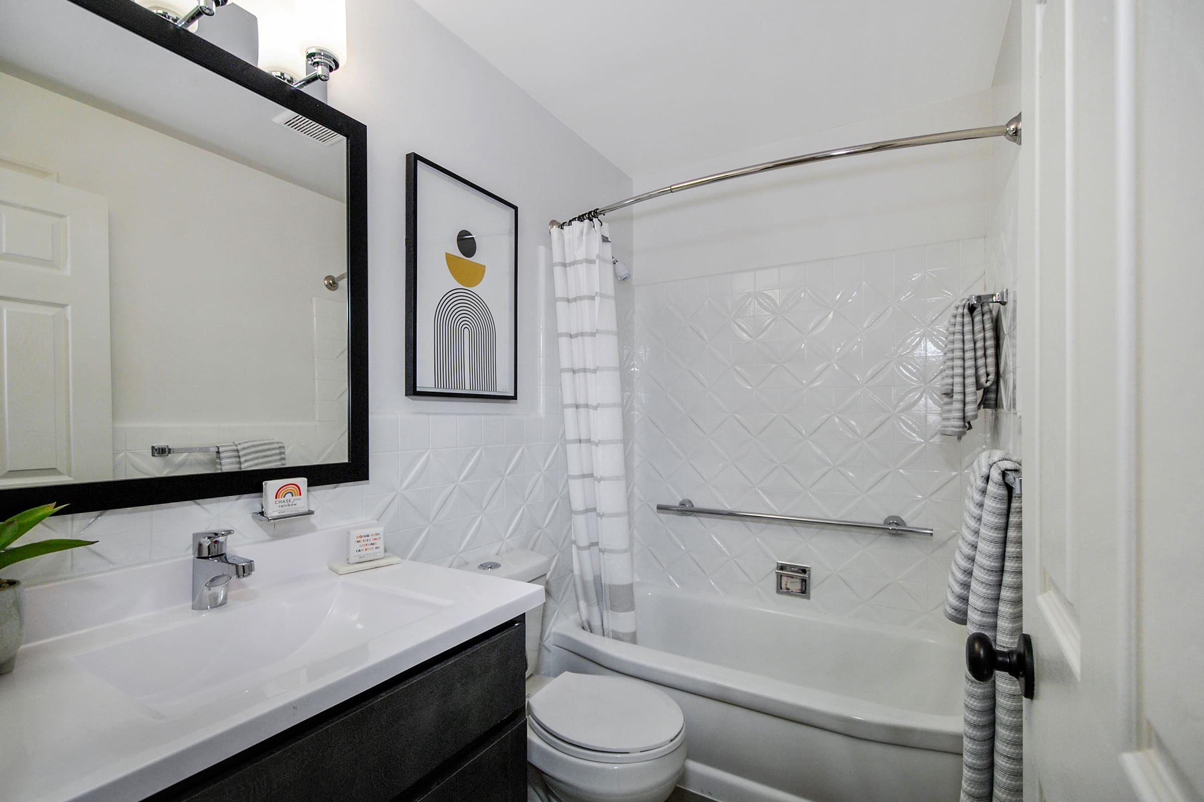 A modern bathroom featuring a white tub with a shower curtain, a large mirror above a sleek sink, and a minimalist design. The walls have a textured tile pattern, and there are gray and black accents, including towels and a potted plant, creating a clean and stylish atmosphere.