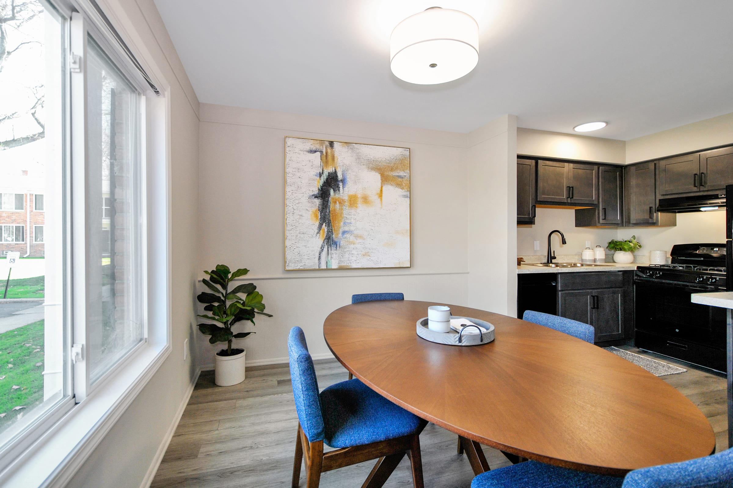 A modern dining area featuring an oval wooden table surrounded by blue upholstered chairs. The room includes a large window with natural light, a potted plant in the corner, and a contemporary abstract painting on the wall above the table. The kitchen is visible in the background with dark cabinets and a stove.