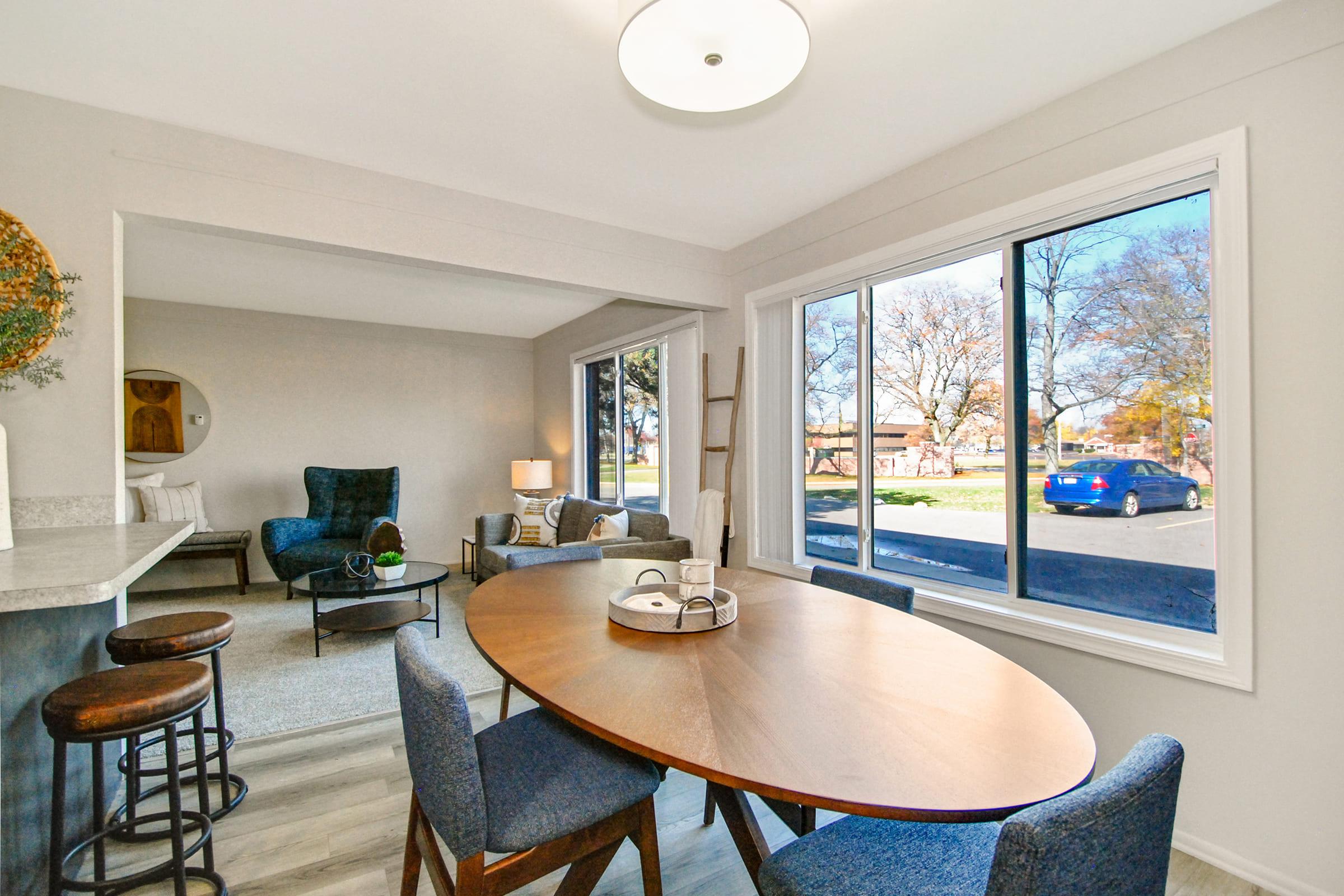 A modern dining area featuring a round wooden table surrounded by blue upholstered chairs. A cozy living space is visible in the background with a blue armchair and lamp. Large windows allow natural light, showcasing a view of trees and a parked blue car outside.