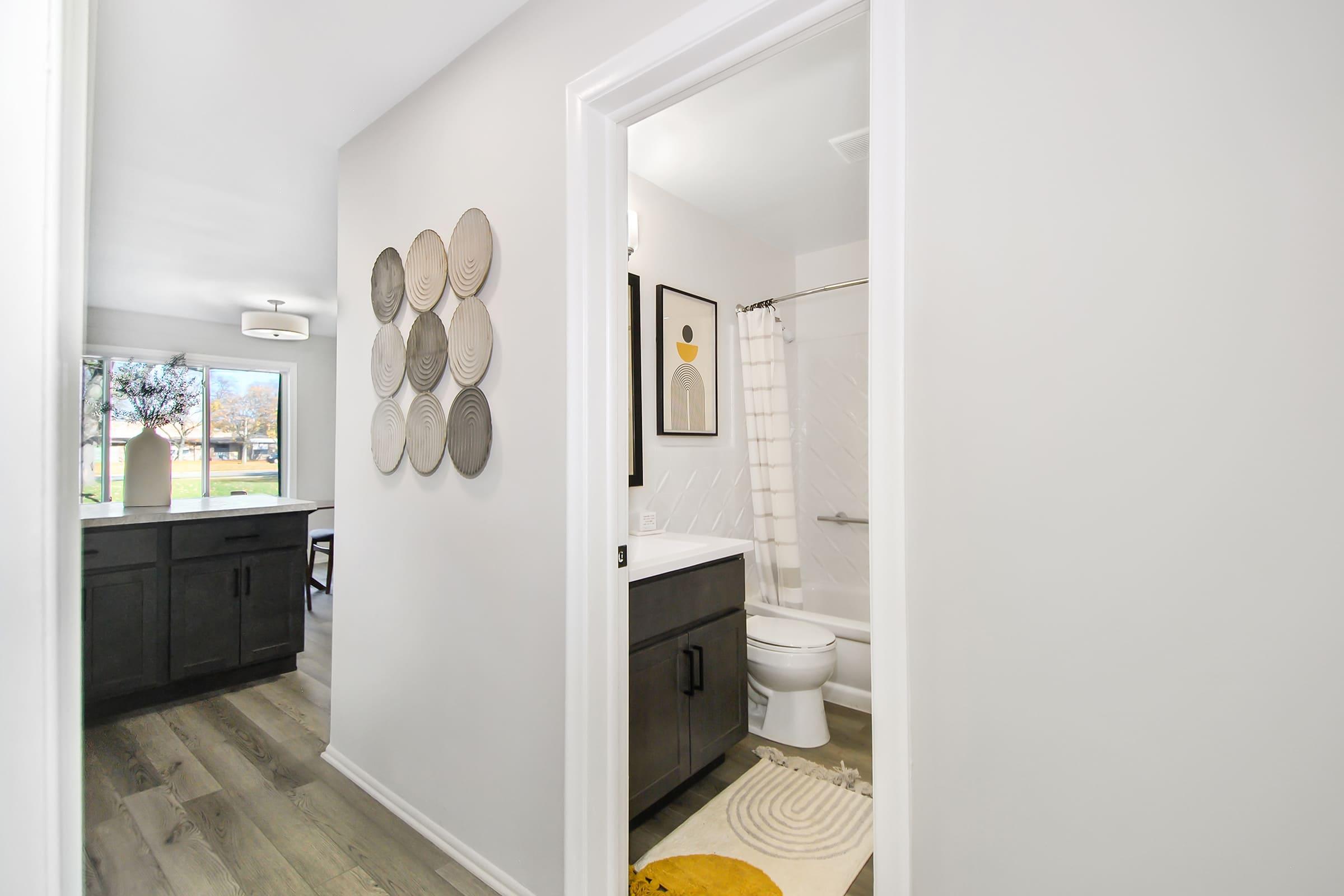 A modern bathroom and adjoining room with light walls, wood flooring, and stylish decor. The bathroom features a shower/tub combination, a toilet, and a sink with dark cabinetry. Decorative plates are hung on the wall. Natural light floods in from a nearby window.