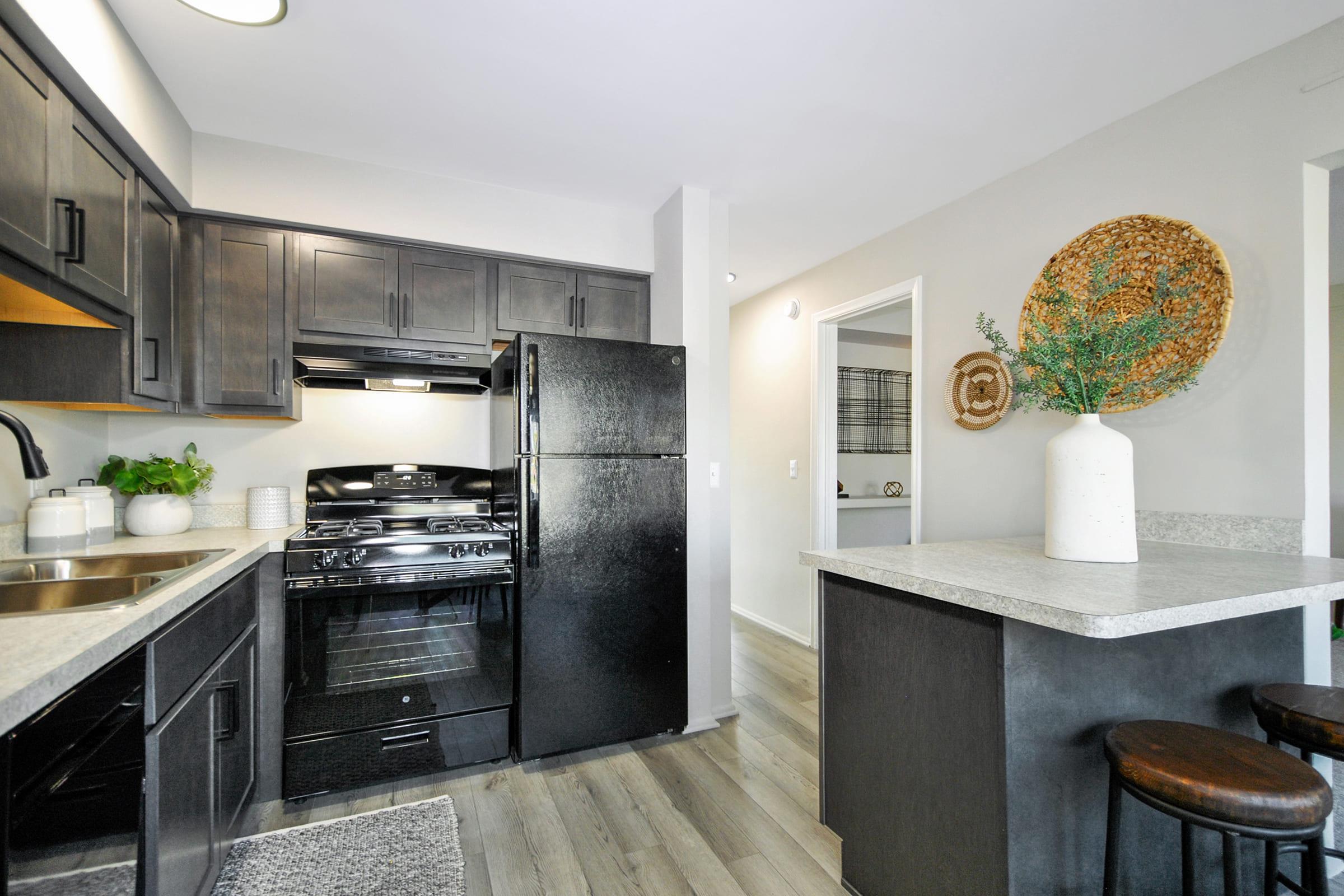 A modern kitchen featuring dark cabinetry, a black refrigerator, and a gas stove. The countertop is light-colored, complementing the floor, which has a wood-like finish. A round woven wall decoration is visible, along with a minimalist vase with greenery on the countertop. Stylish bar stools are positioned at the counter.