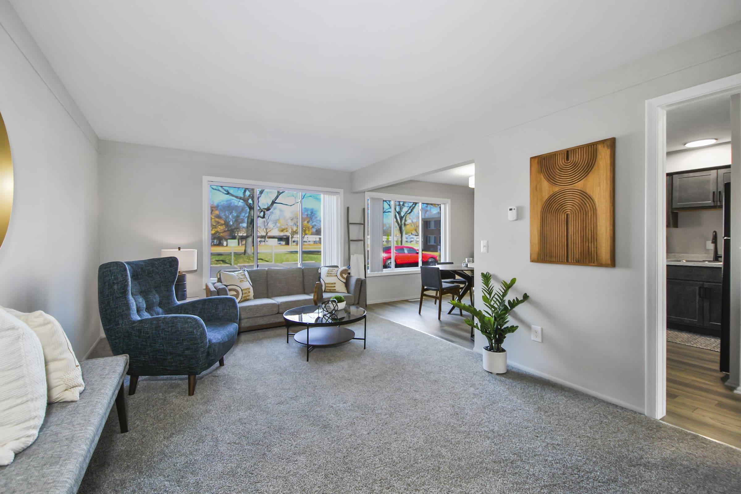 A modern living room featuring a cozy seating area with a blue armchair and a gray sofa, a small round coffee table, and a potted plant. Large windows allow natural light to fill the space, showcasing a view of trees and a red car outside. The walls are painted light gray with decorative elements.