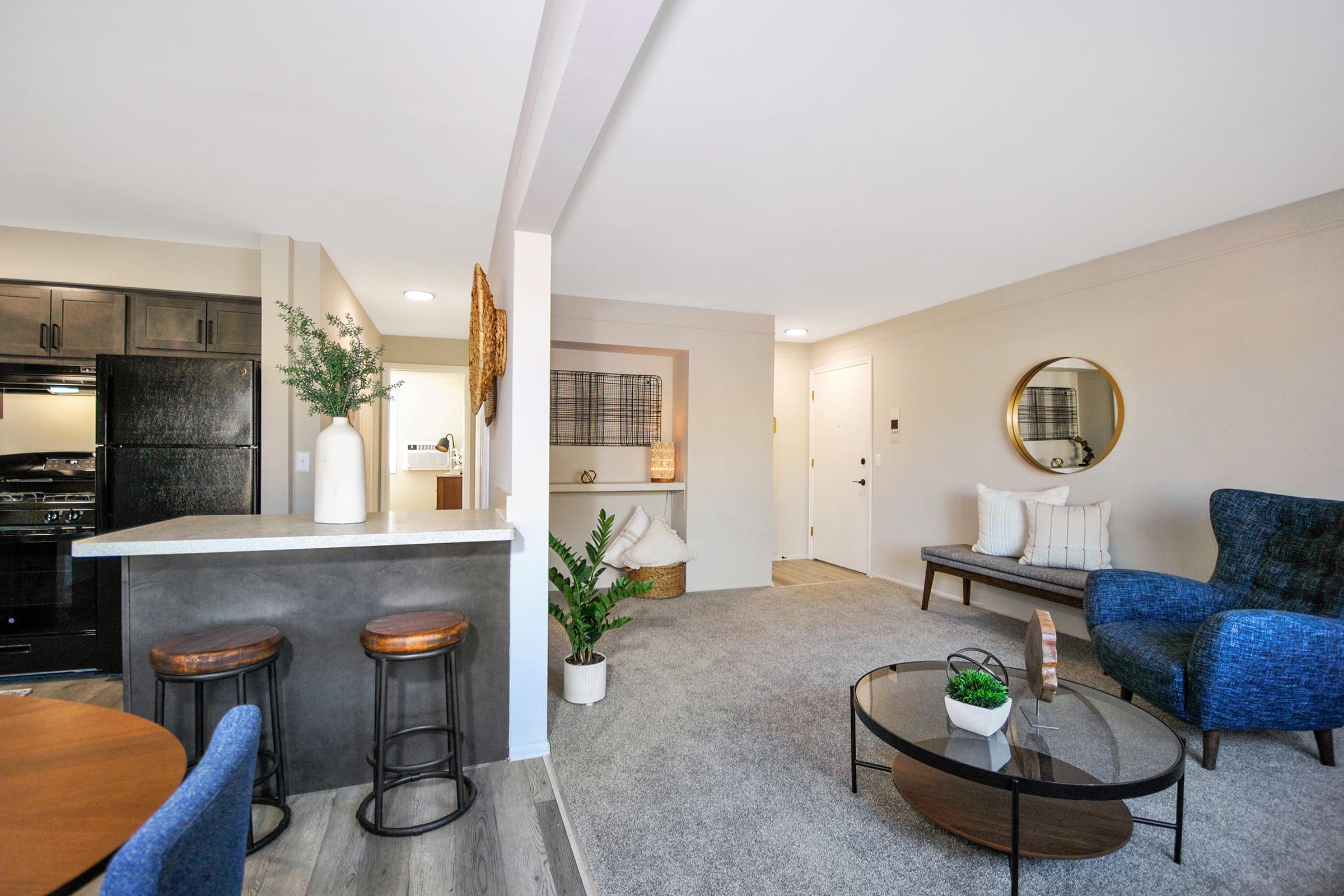 A modern living space featuring a cozy seating area with a blue armchair, a round coffee table, and a neutral-colored rug. The kitchen bar area, with dark cabinetry, is visible on one side. A decorative plant adds a touch of greenery, while a round mirror and wall art enhance the aesthetic of the room.