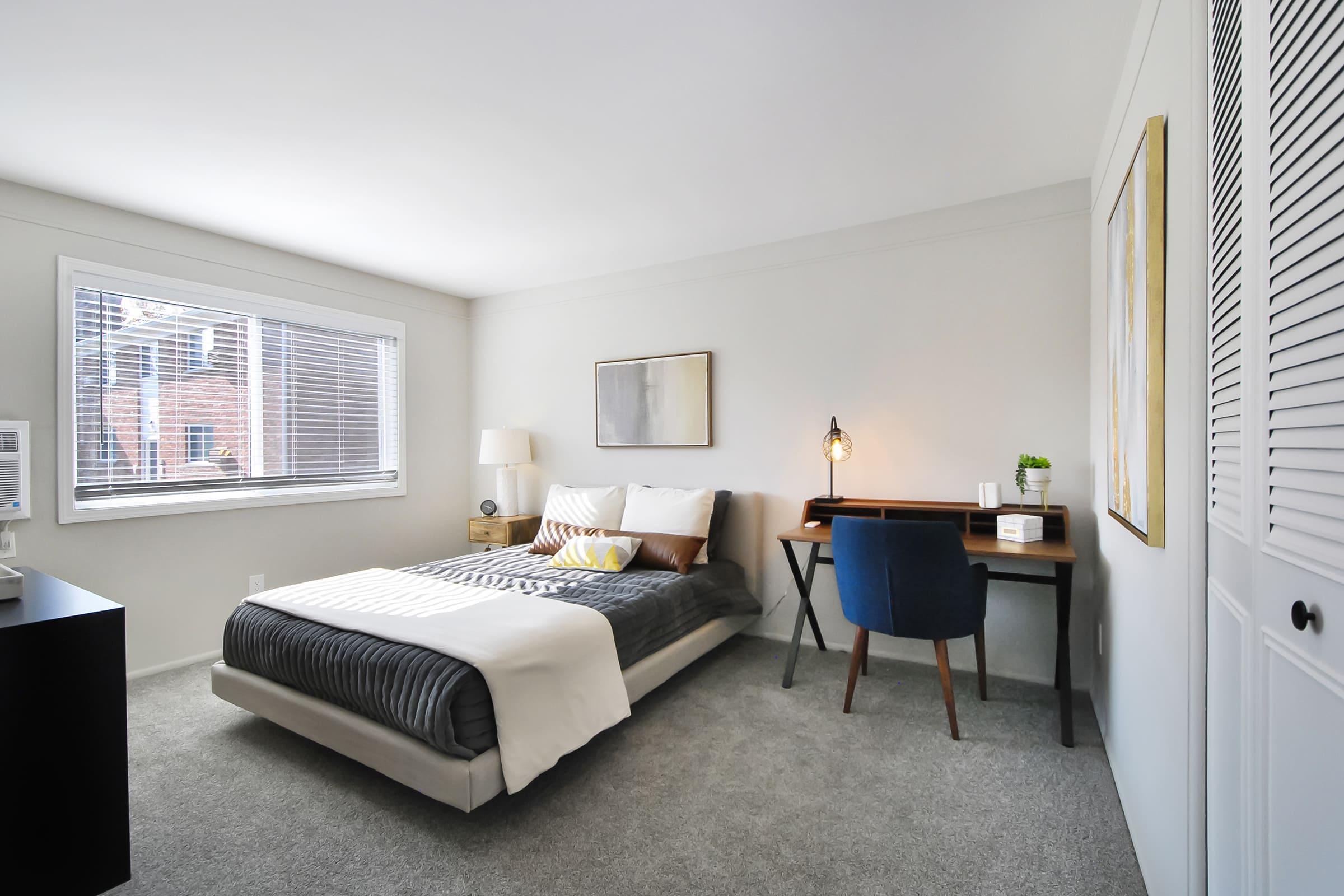 A well-lit contemporary bedroom featuring a cozy bed with decorative pillows, a bedside table with a lamp, and a stylish desk with a chair. The room has light-colored walls, a window with city views, and a soft carpet, creating a warm and inviting atmosphere.