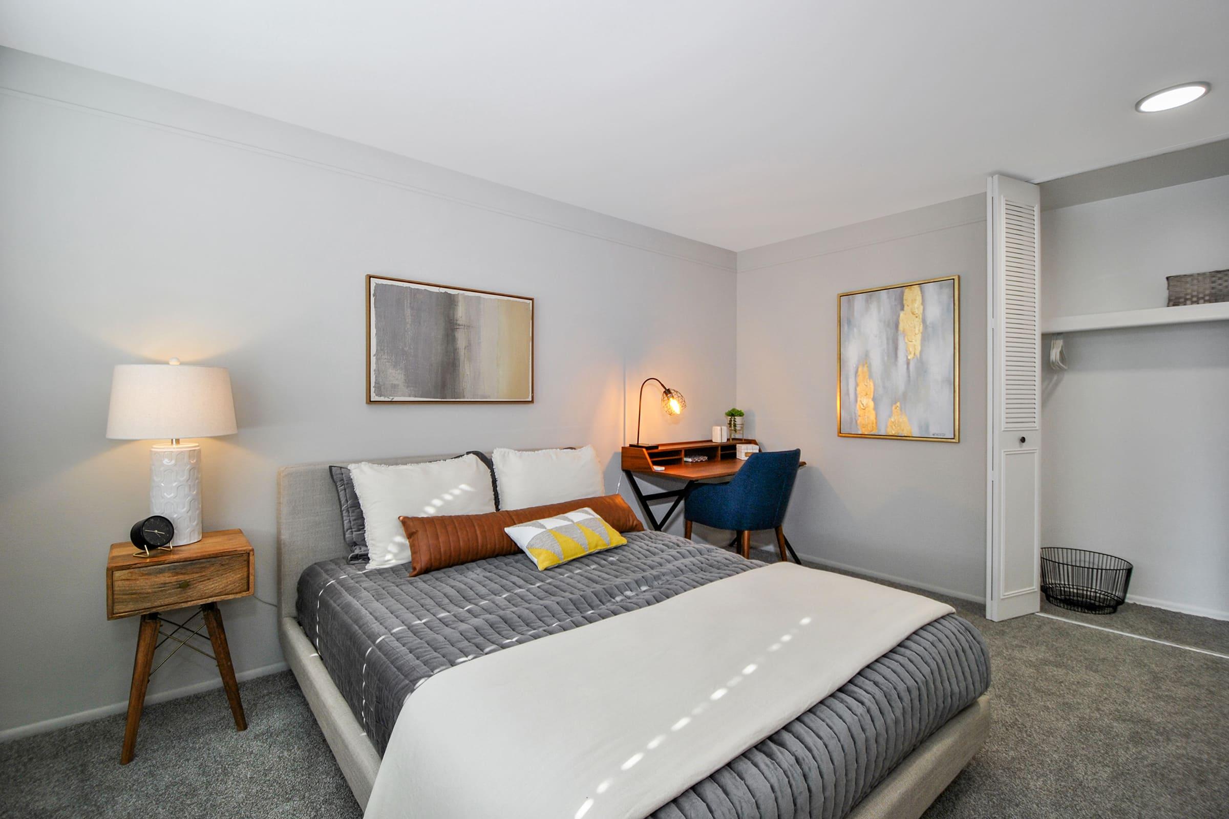 A modern bedroom featuring a comfortable bed with gray bedding and a decorative pillow. There's a nightstand with a lamp, a small desk with a blue chair, and two framed artworks on the walls. The room has soft lighting and neutral-colored walls, creating a cozy atmosphere. A small shelf and a basket are also visible.