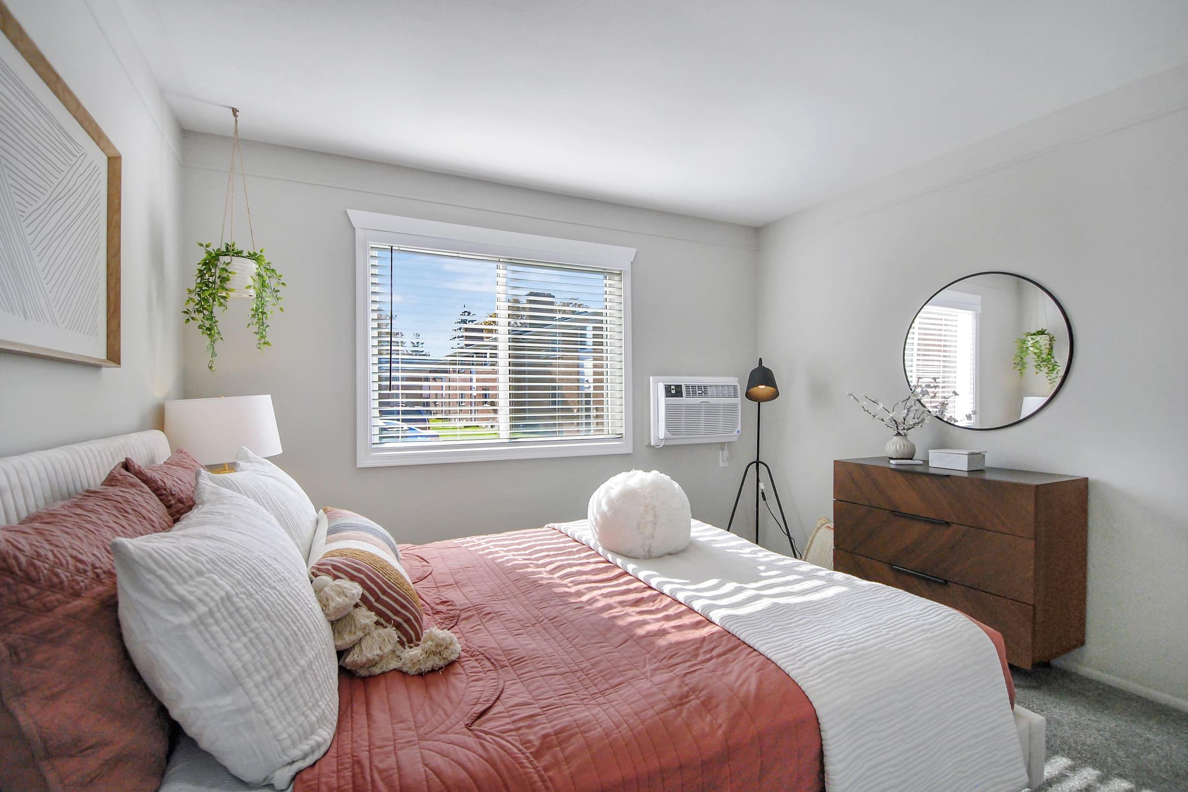 A cozy bedroom featuring a bed with a pink and white comforter, decorative pillows, and a fluffy throw. There's a round mirror on the wall, a lamp beside the bed, and a dresser. A window allows natural light to brighten the room, with greenery hanging nearby and a landscape visible outside.