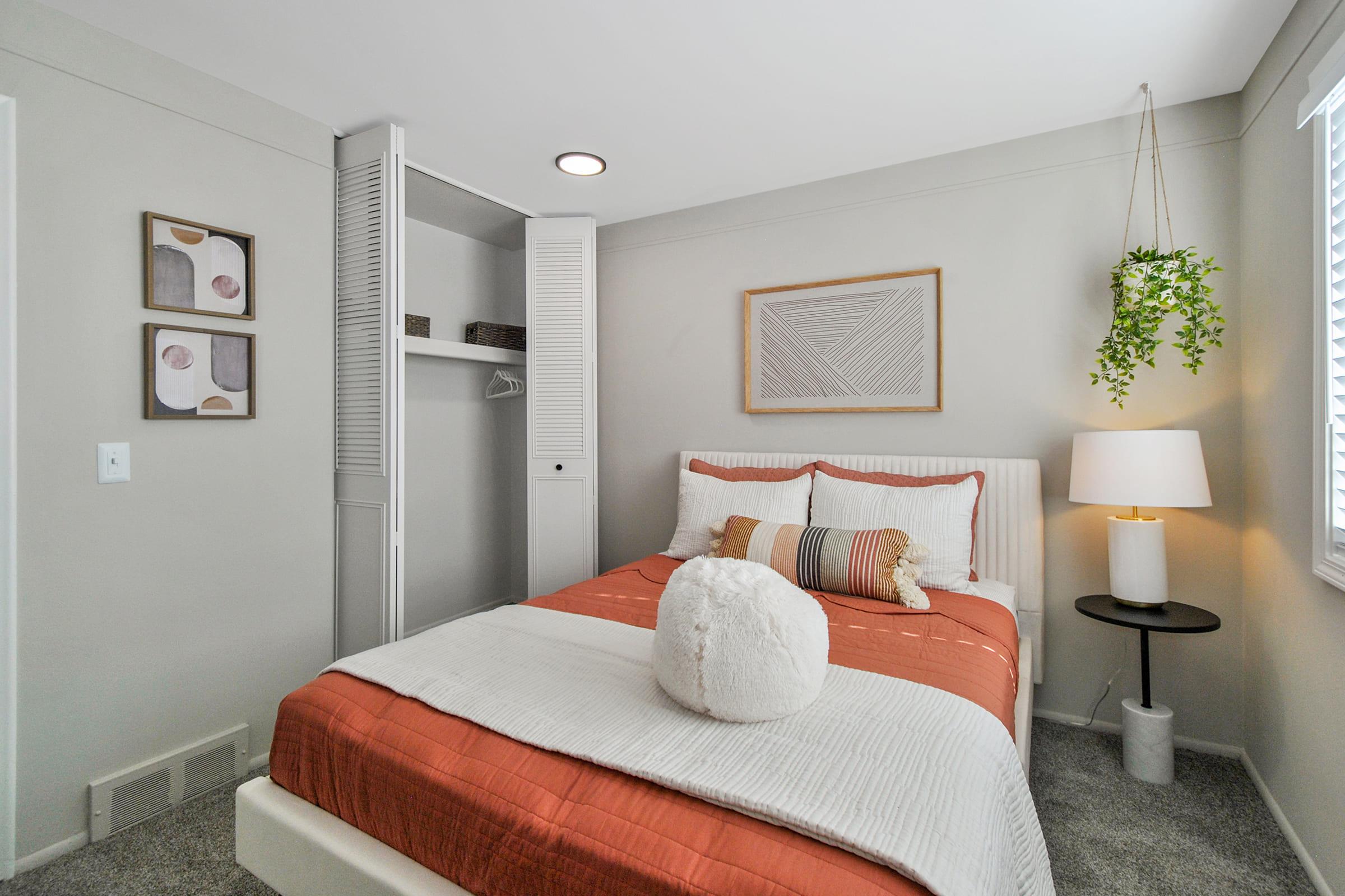 A cozy bedroom featuring a soft orange and white bedspread, fluffy white pillow, and minimalist decor. There's a small nightstand with a lamp, framed art on the walls, and a closet space with sliding doors. A hanging plant adds a touch of greenery, and natural light streams in through a window.