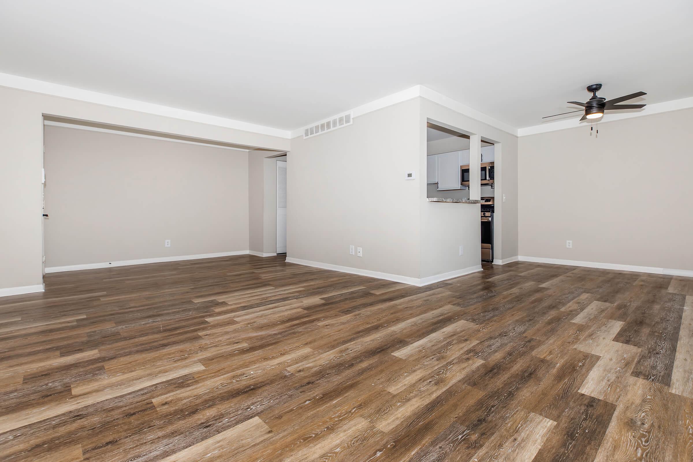 An empty, well-lit room with light beige walls and wooden laminate flooring. A ceiling fan is mounted near the center, and a doorway leads to another room. In the background, there is a glimpse of a kitchen area with modern cabinetry. The space is clean and unfurnished, giving a sense of openness.