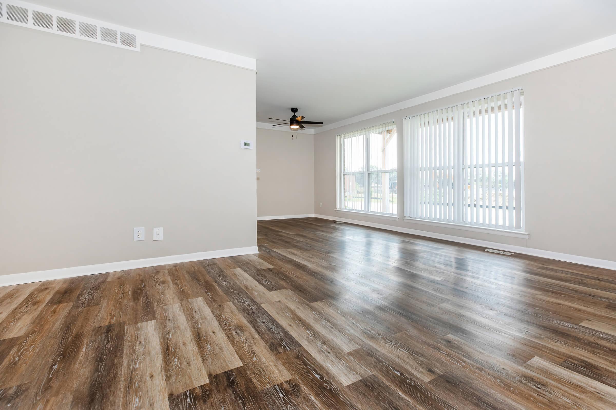 Spacious, well-lit living room with large windows featuring vertical blinds. The room has a modern ceiling fan and stylish wood-like flooring, creating a warm and inviting atmosphere. The walls are painted a light neutral color, enhancing the sense of openness.