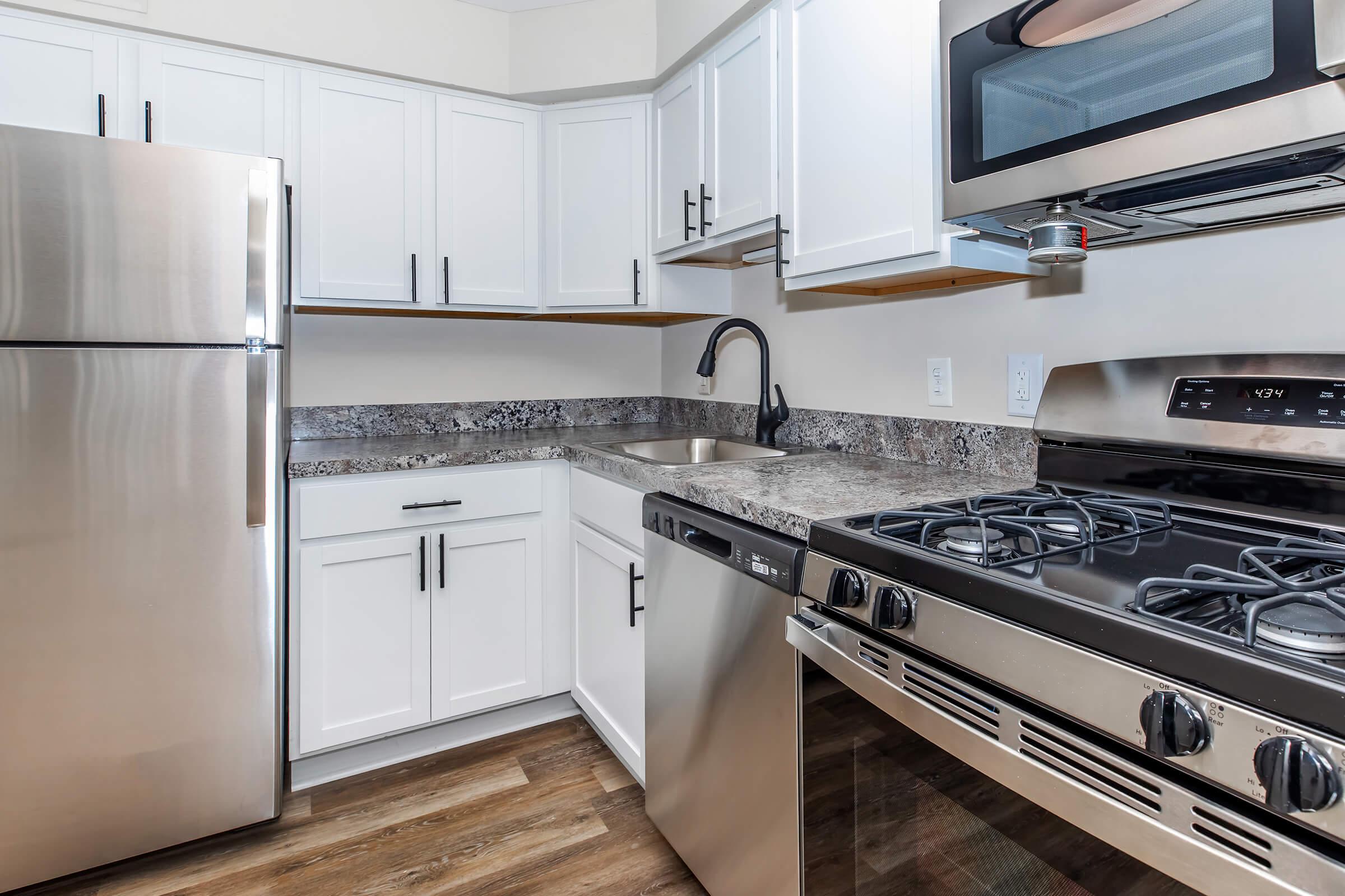 Modern kitchen featuring stainless steel appliances, including a refrigerator, dishwasher, and gas stove. The countertops are dark granite, and the cabinets are white with sleek handles. A black faucet is installed above the sink, and the flooring is a warm-toned wood laminate, creating a stylish and functional cooking space.