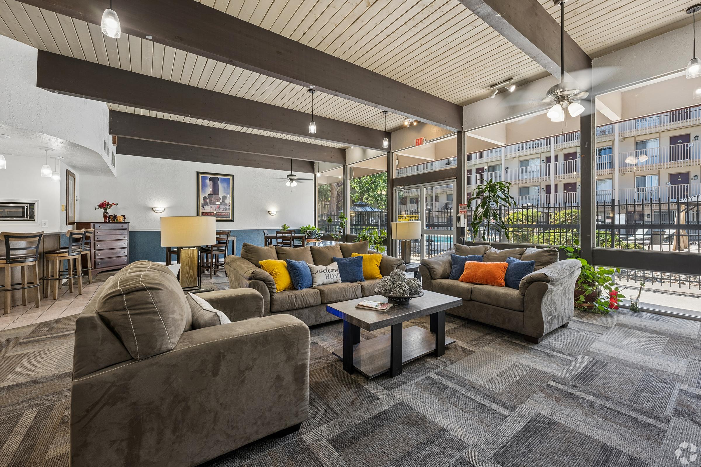 A comfortable lounge area featuring large sofas adorned with colorful throw pillows, a coffee table, and potted plants. Large windows allow natural light to fill the space, which includes a dining area in the background with wooden tables and chairs. The decor is modern and inviting, ideal for relaxation.