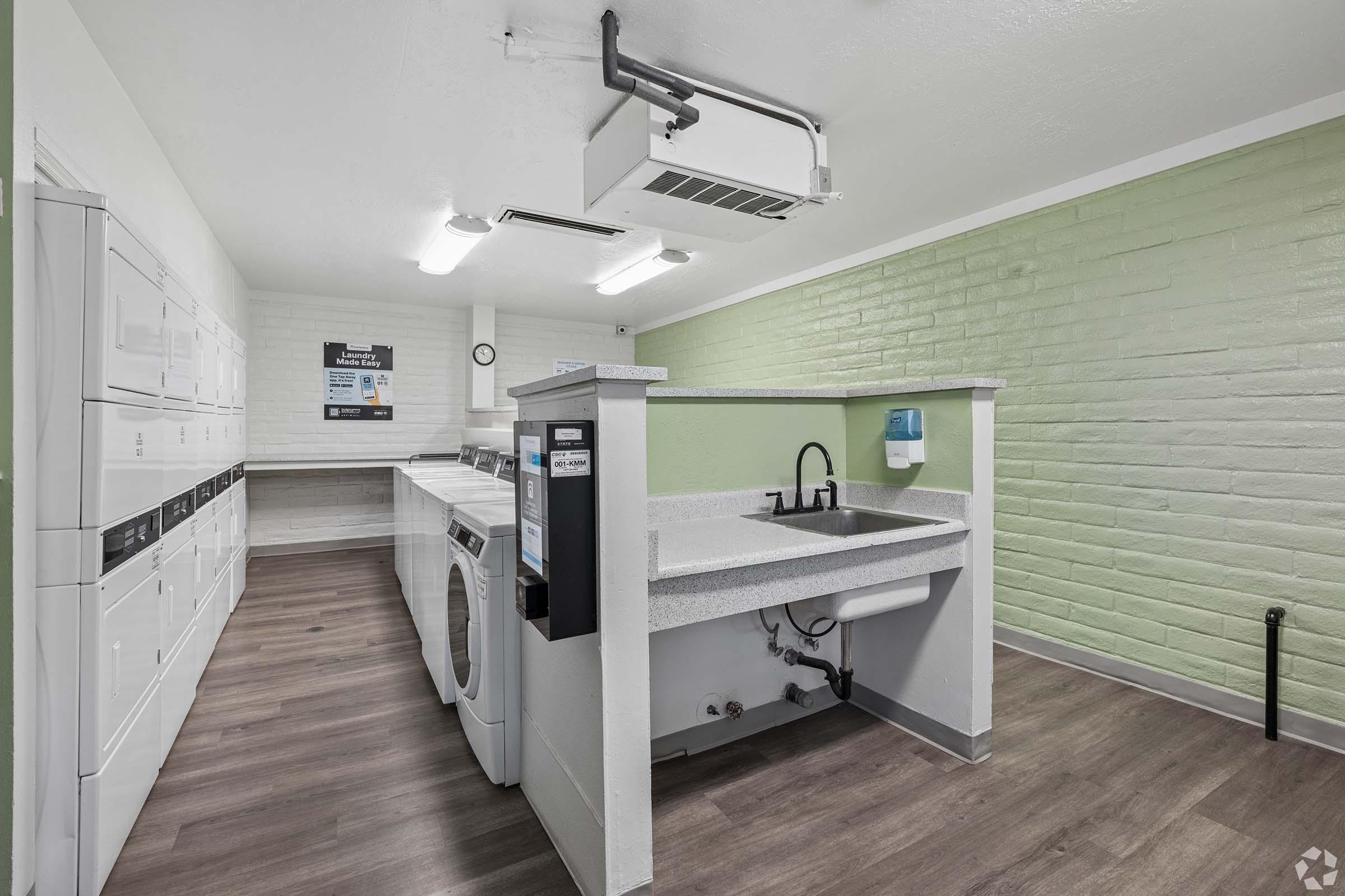 A clean and modern laundry room featuring multiple washing machines and dryers lined against the wall. There is a utility sink with a countertop in the center, and light green painted walls paired with light wood-like flooring create a bright atmosphere. A clock is mounted on the wall for convenience.